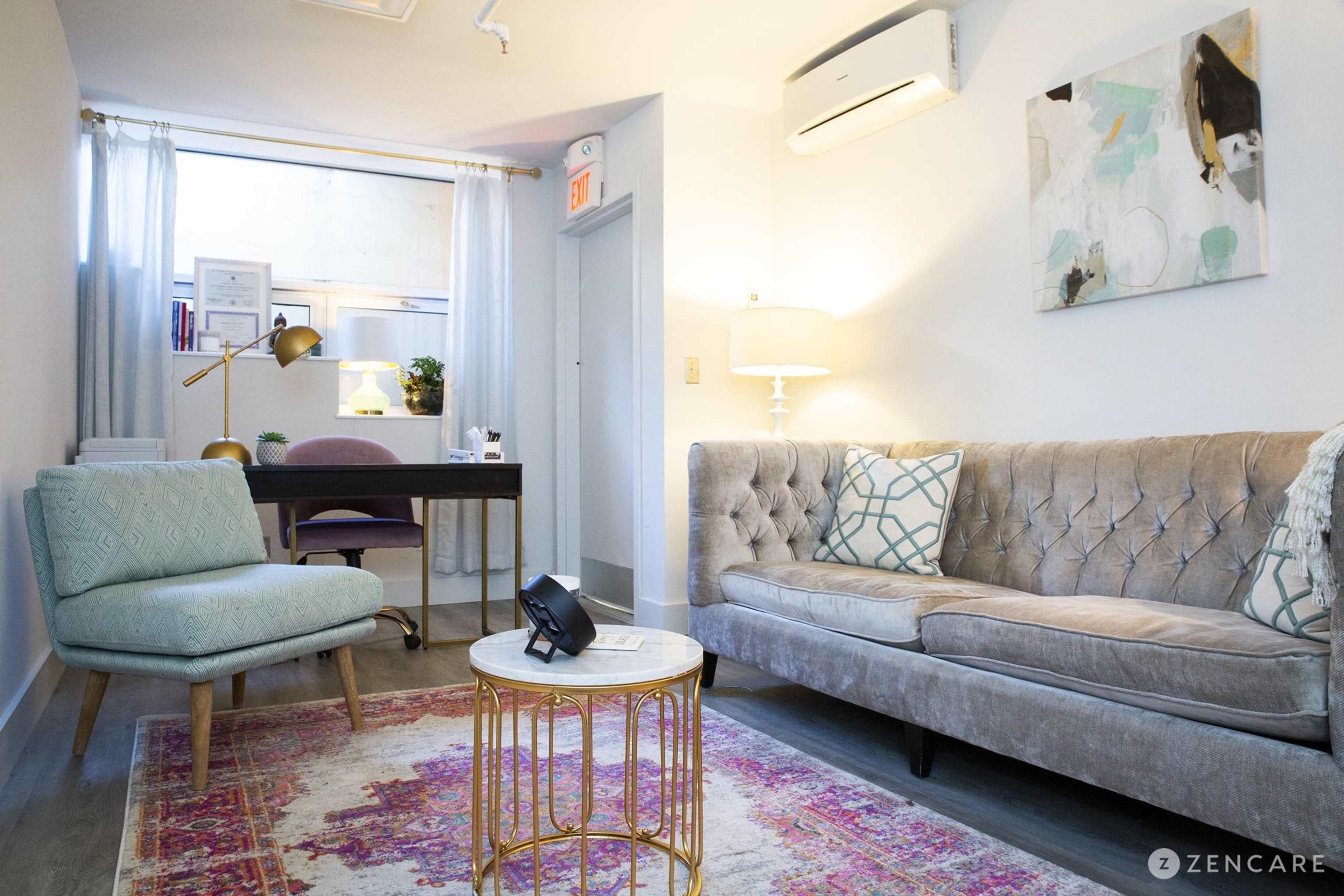 The image shows a modern room with a tufted sofa, a small round table, a light blue chair, and a desk in the background under a window.