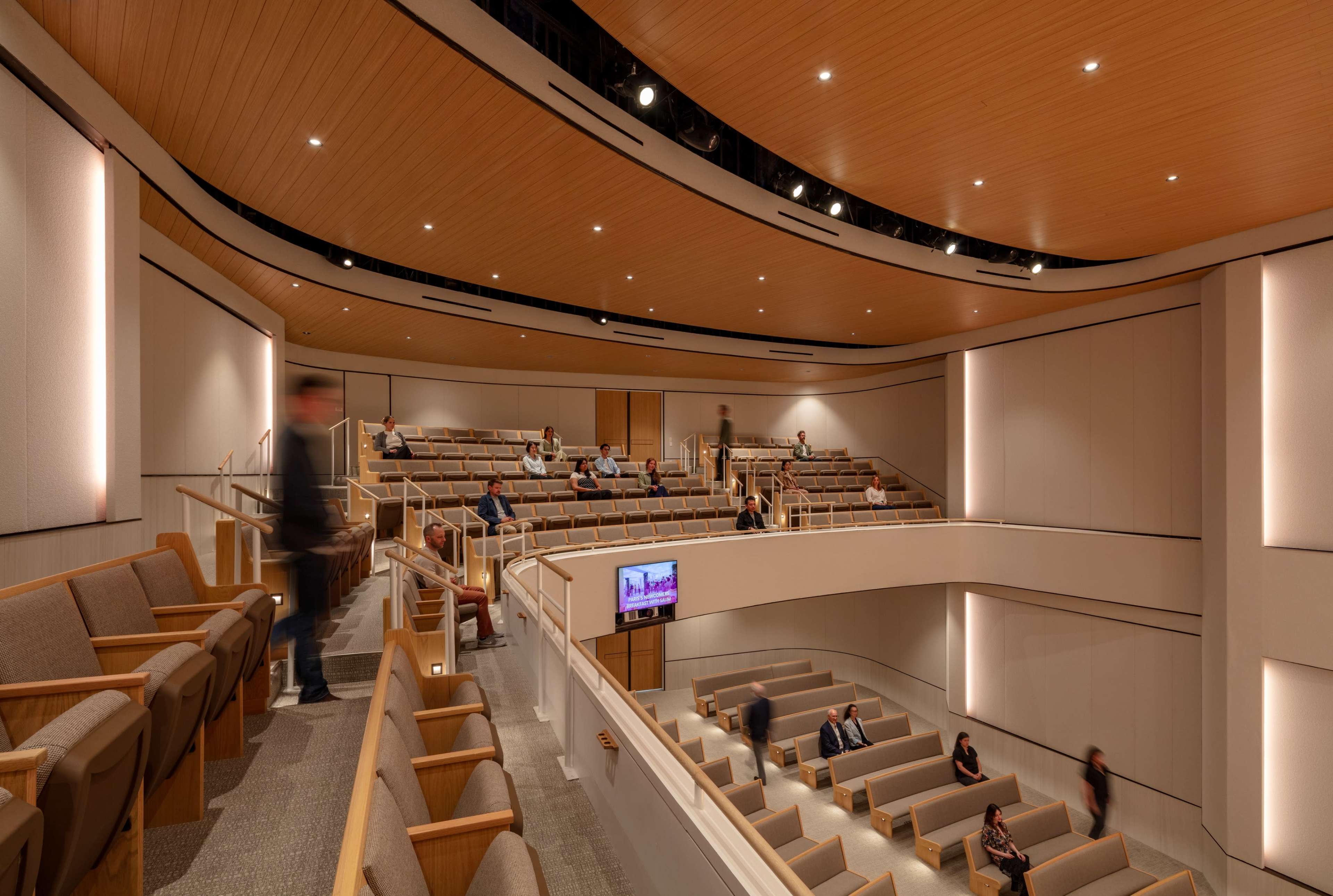 The image shows an interior of a modern auditorium with tiered seating and a wooden ceiling, partially filled with people.