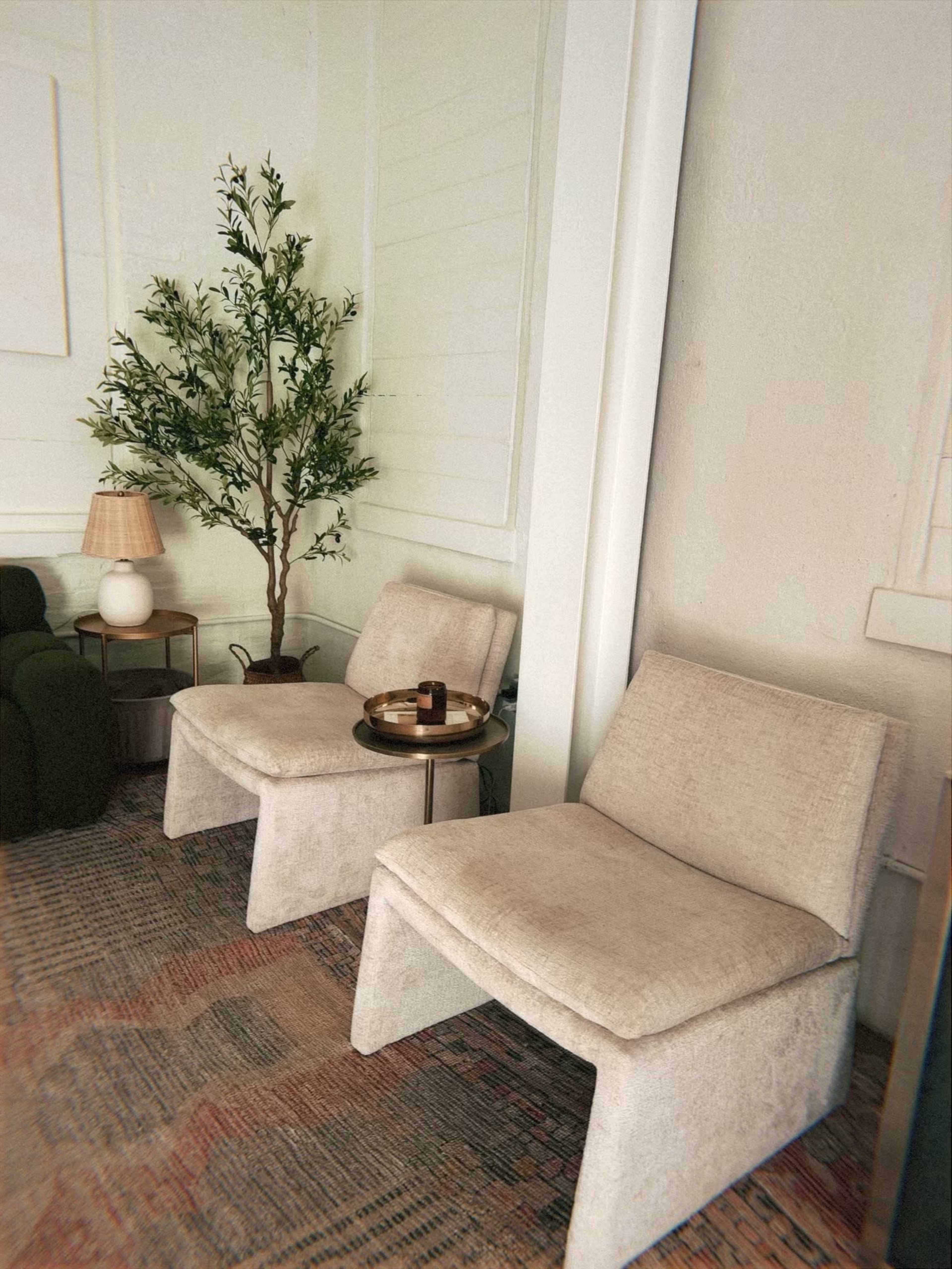 Two beige chairs are positioned beside a small round table with a decorative item, while a potted plant and a lamp rest nearby in a modern interior setting.