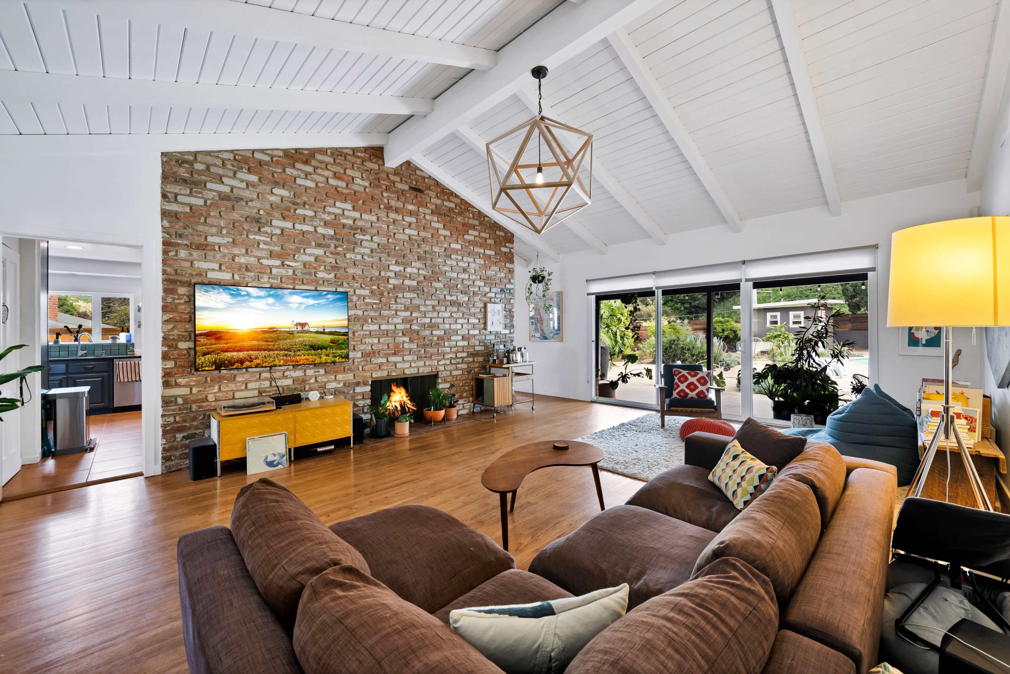 The image shows a spacious living room with a large brick fireplace, a television mounted on the wall, and a sectional couch, all illuminated by natural light from large windows.