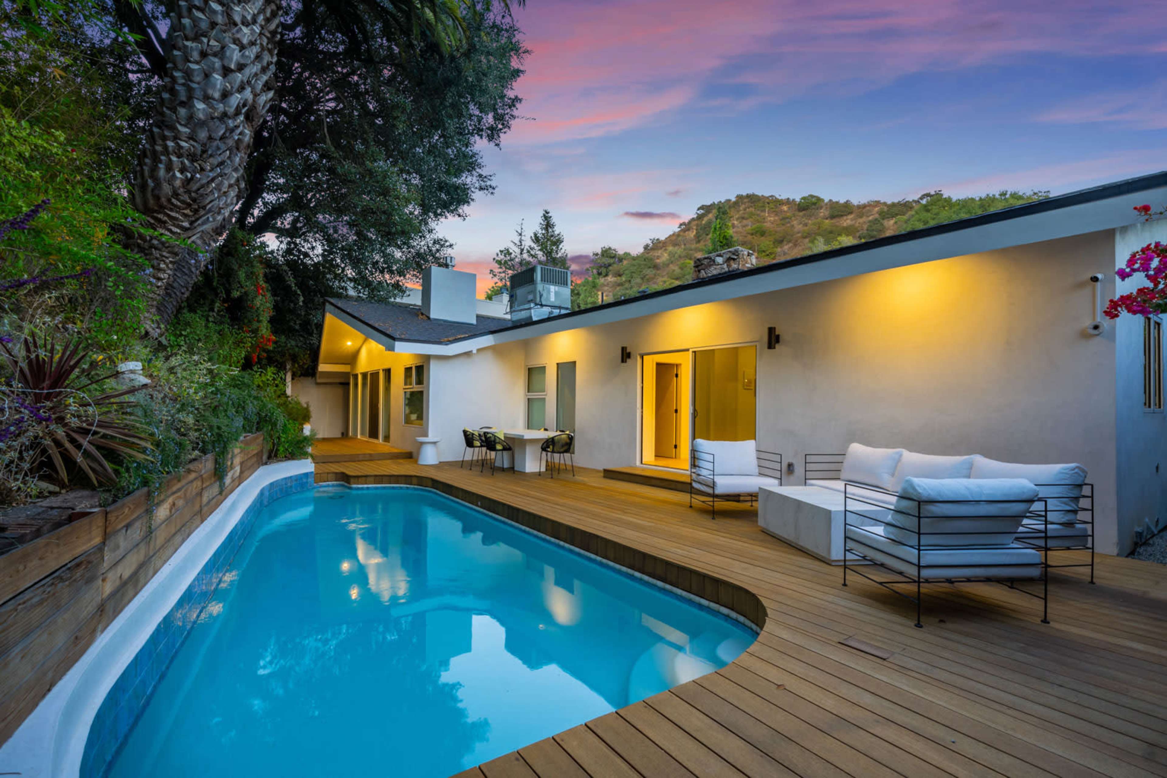 A modern house with a swimming pool and wooden deck is set against a hillside backdrop under a colorful sky.