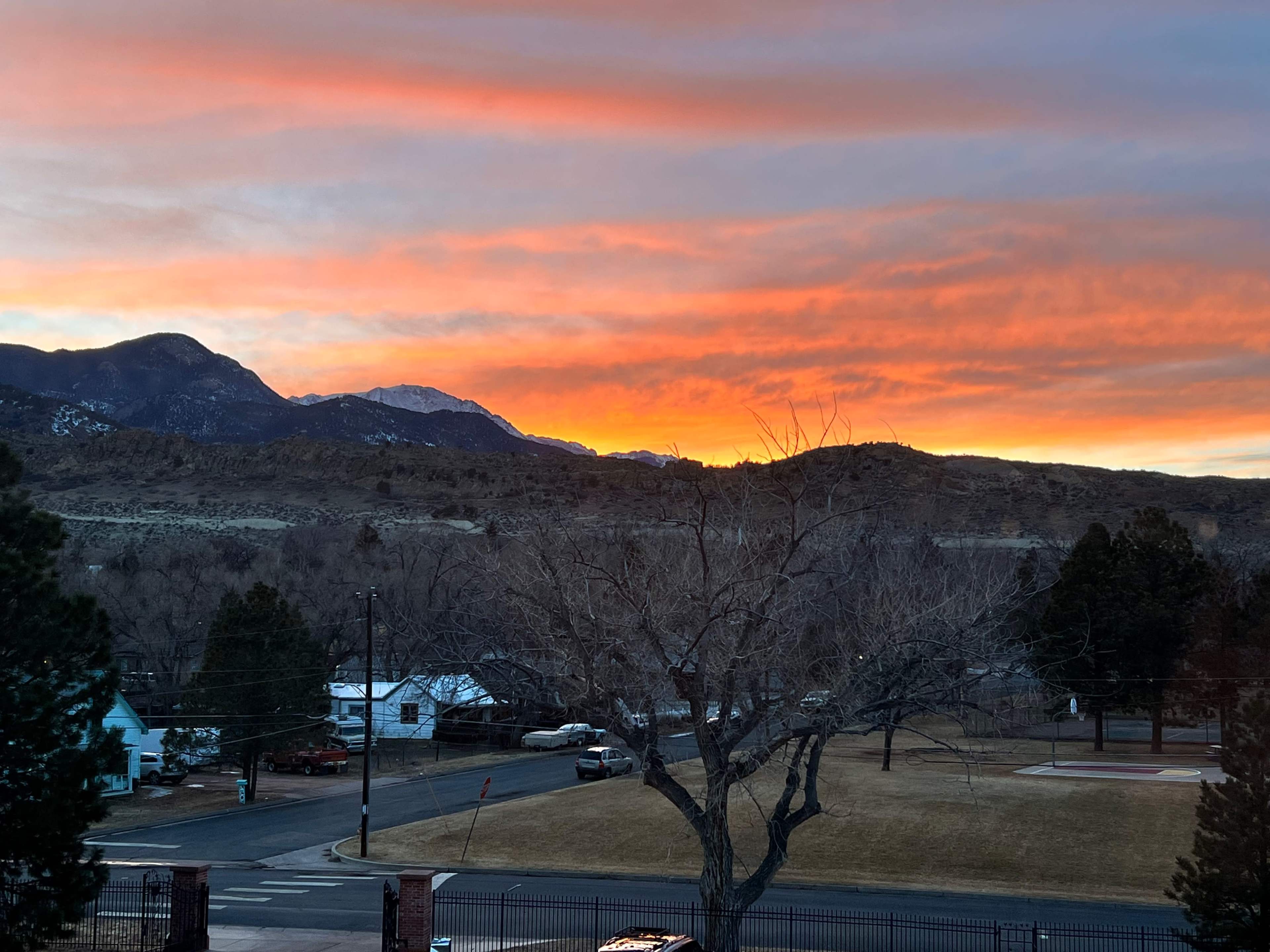 A sunset with vibrant orange and pink hues over mountains, viewed from a residential area with bare trees and a street in the foreground.