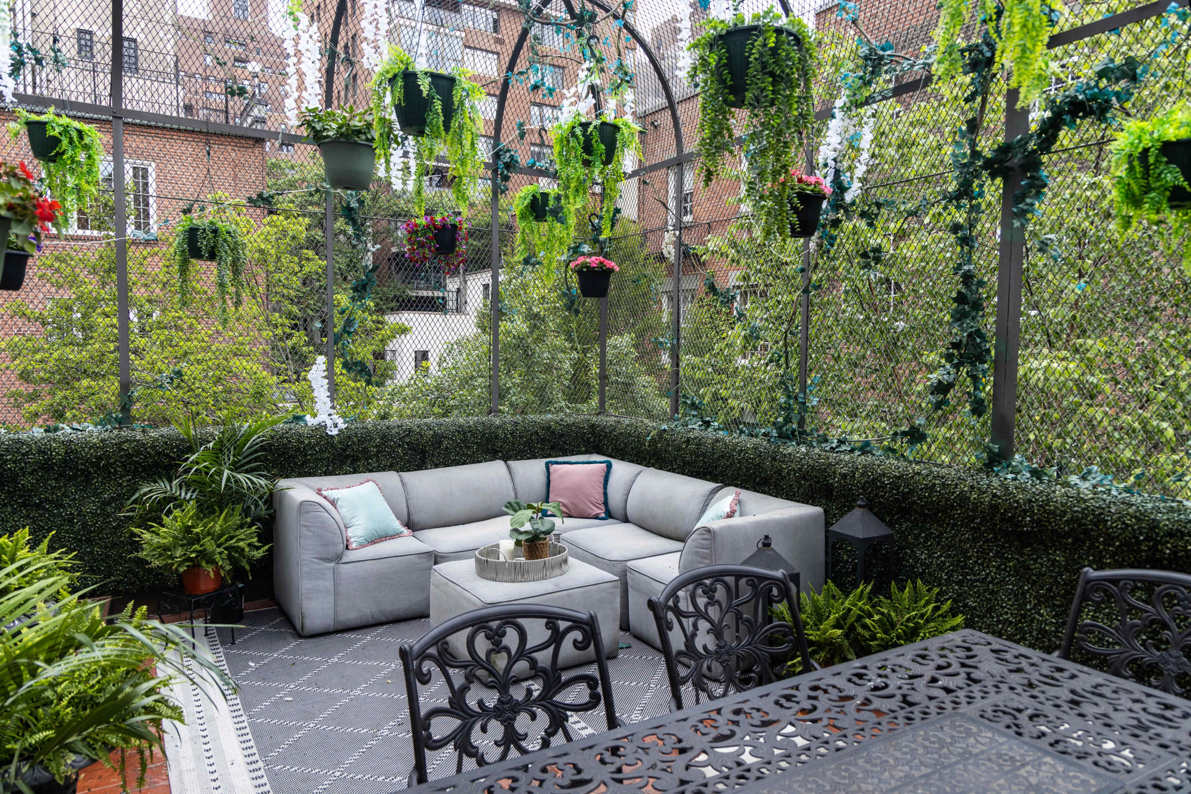 A modern outdoor seating area is surrounded by greenery and hanging plants, featuring a gray sectional couch and a black dining table.