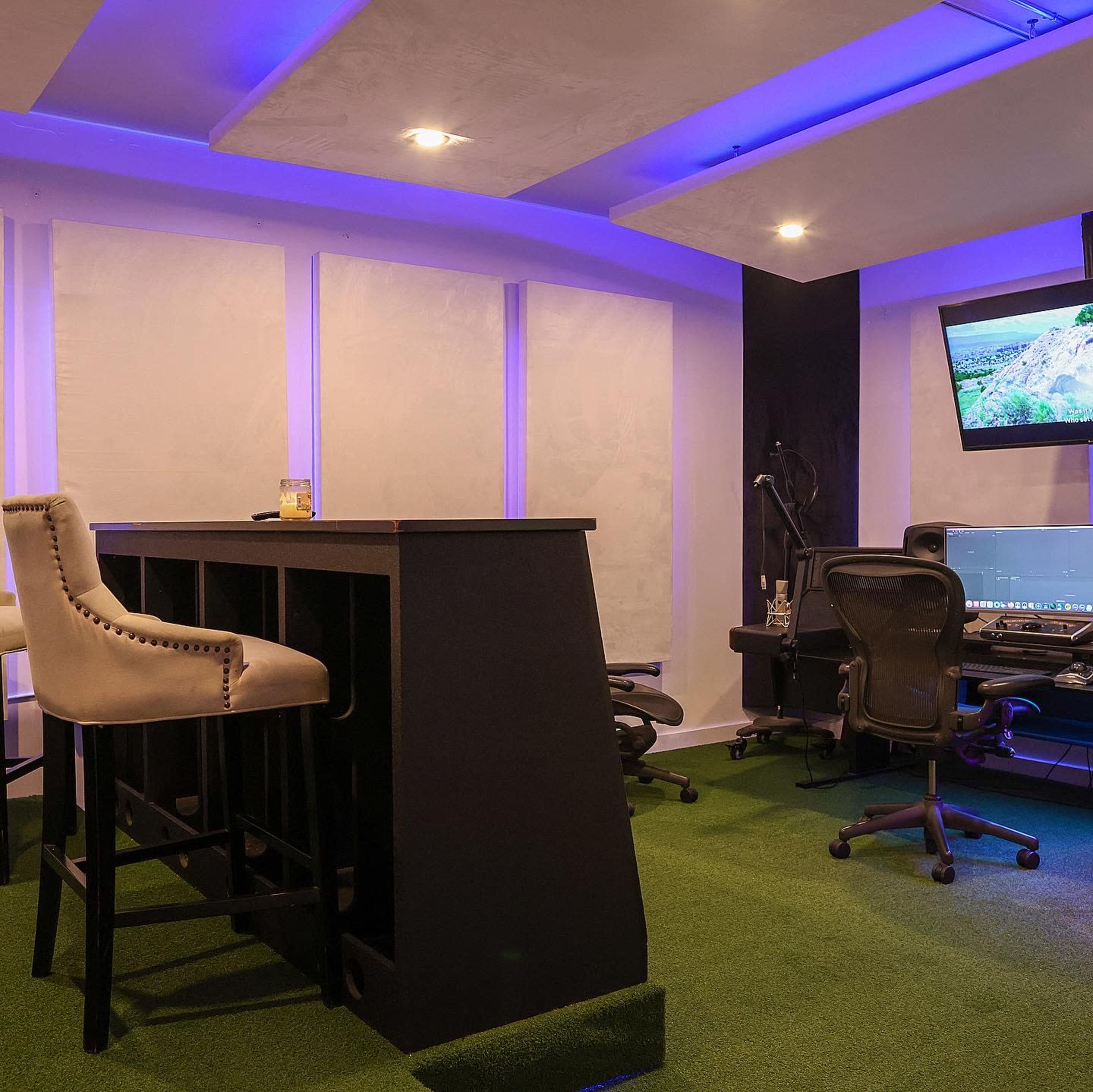 The image shows a modern room with a green carpet, featuring a bar-style counter, office chairs, and a monitor displaying a landscape.