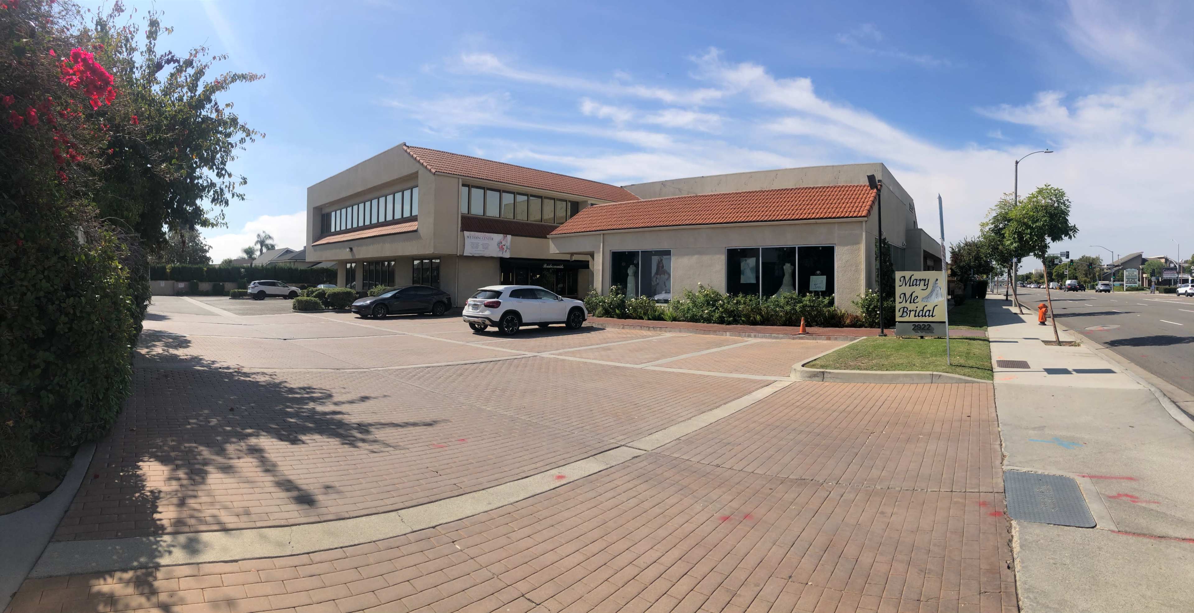 A two-story commercial building with a red-tiled roof, surrounded by a paved parking area and blooming plants.