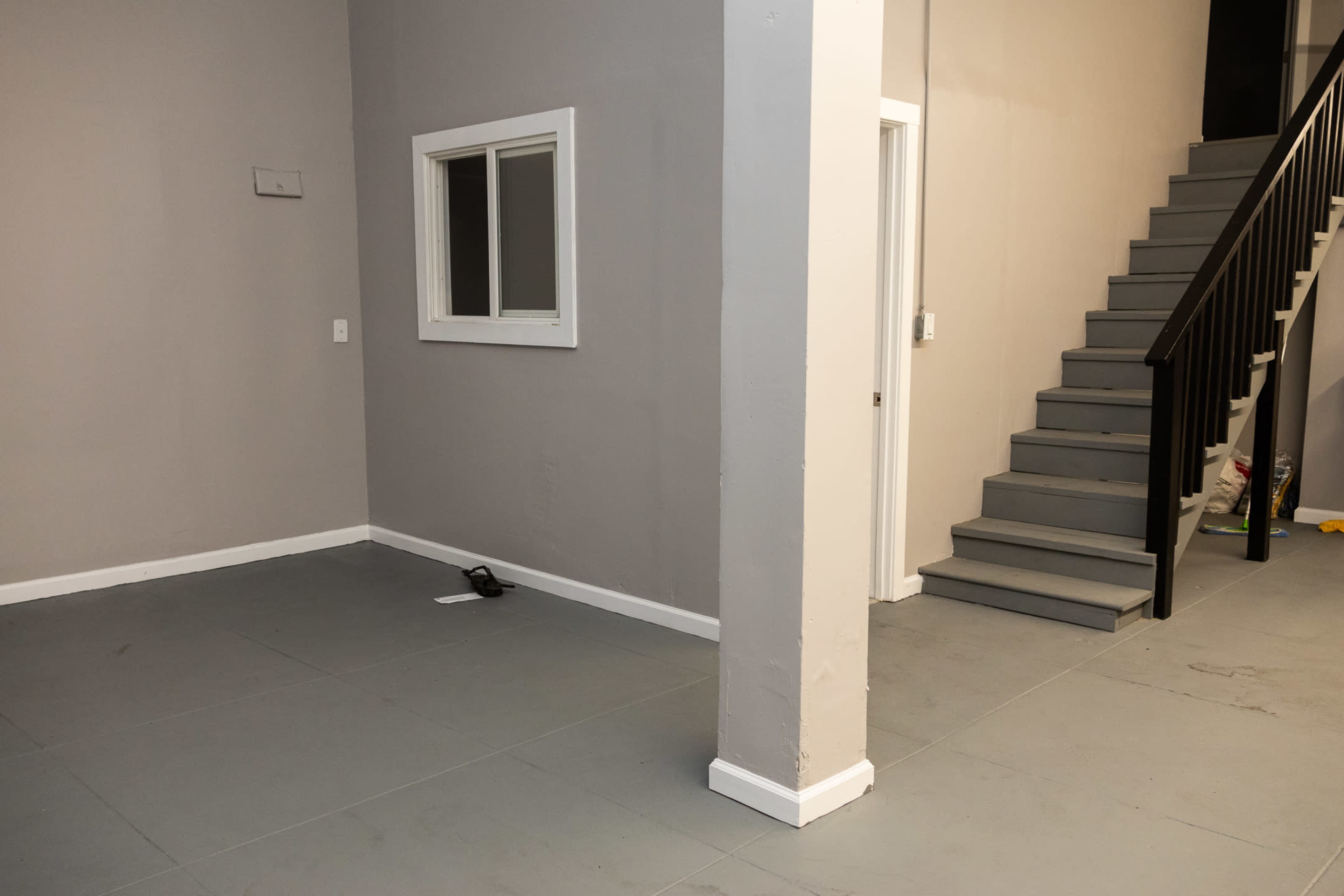 The image shows an interior space with gray walls, a staircase on the right leading to a second level, and a small window cut into the wall on the left.