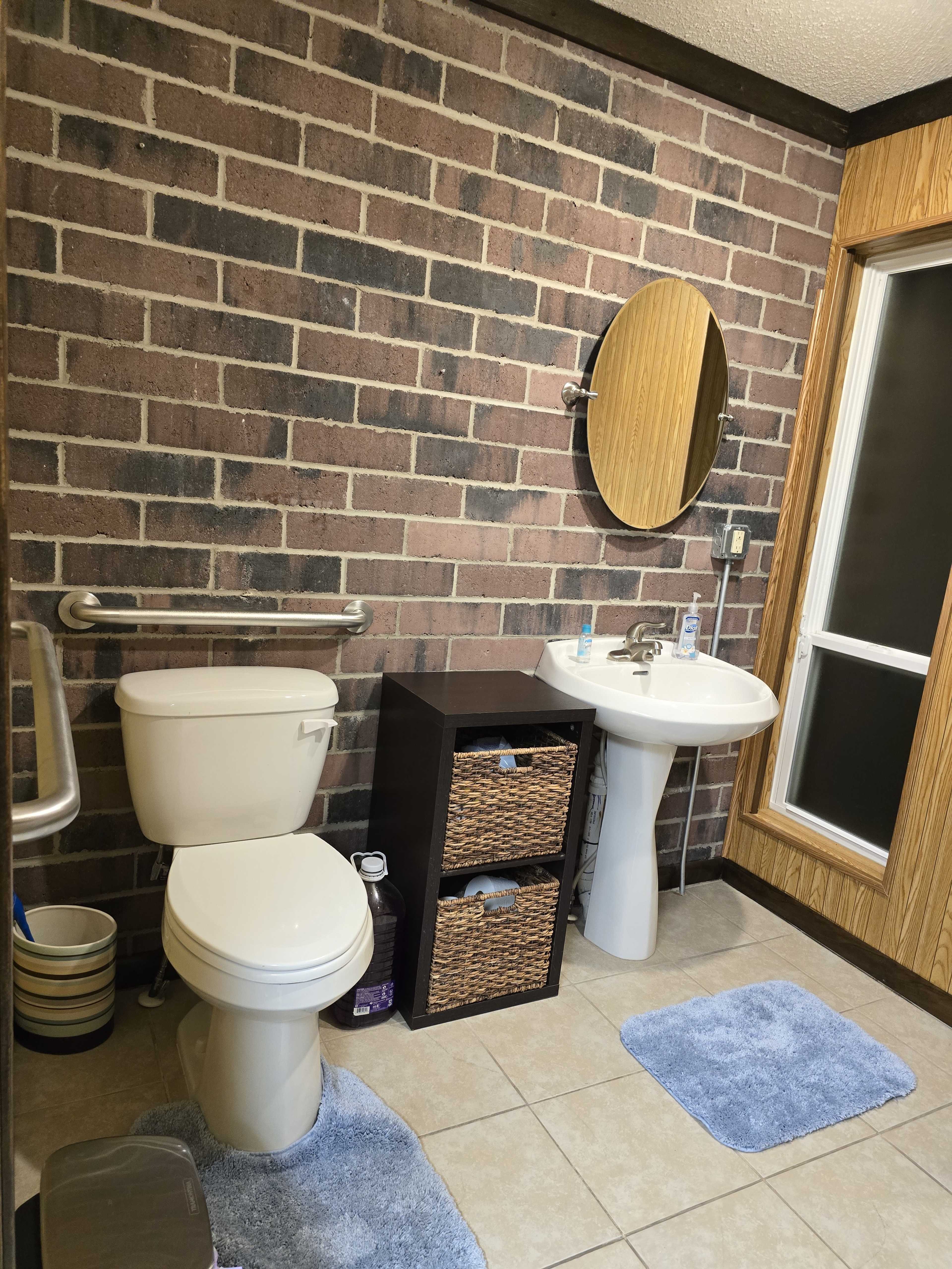 The image shows a bathroom with a toilet, a sink with a mirror, and a storage unit, all set against a backdrop of exposed brick walls.