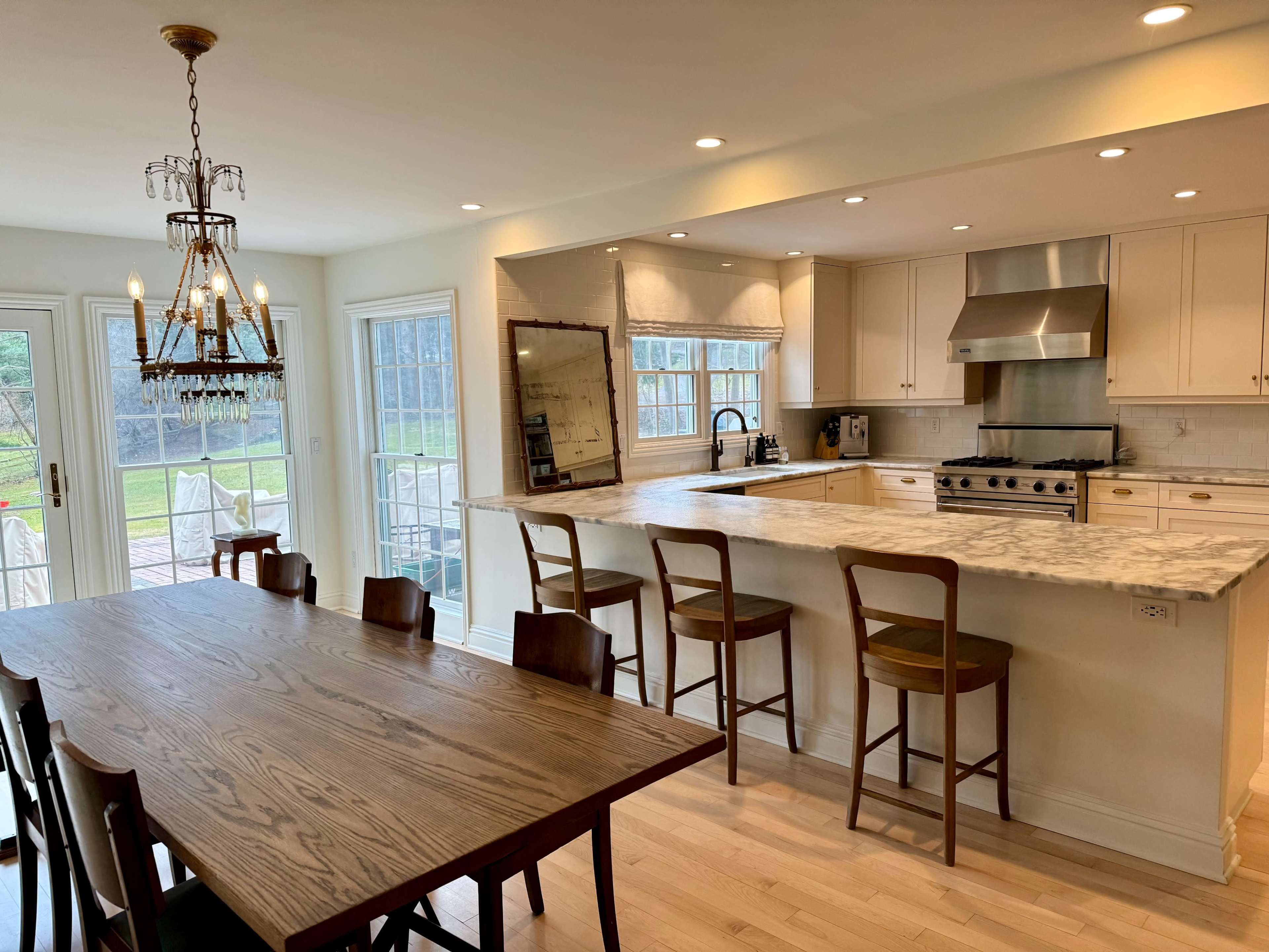 The image shows a modern kitchen and dining area with a wooden table, high-backed chairs, and a spacious countertop next to large windows.