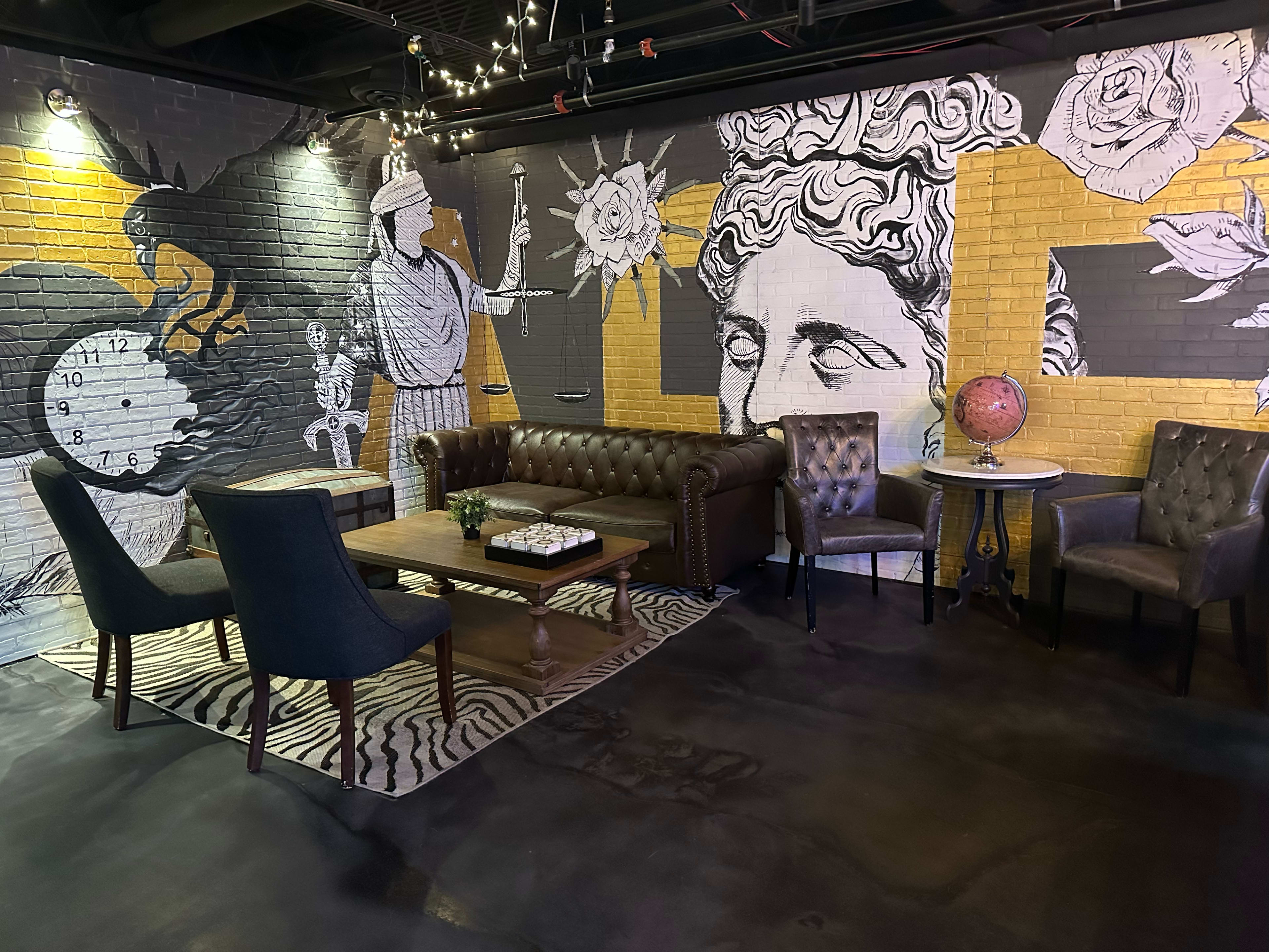 The image shows a lounge area featuring a brown leather couch, two armchairs, a coffee table, and a wall mural with various artistic elements including a clock, a globe, and classical figures.