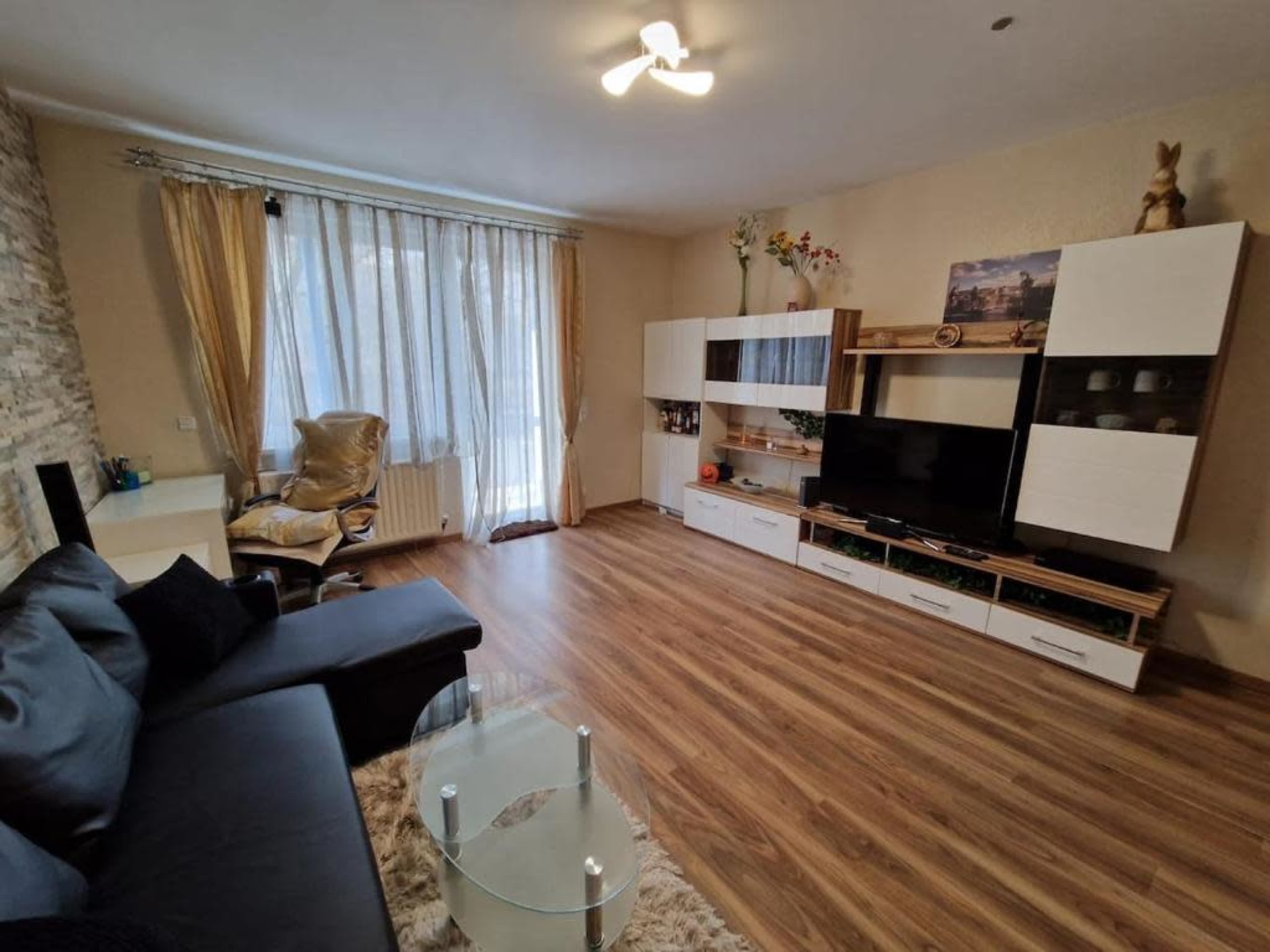 The image depicts a modern living room with a large window, a black sofa, a glass coffee table, and a white entertainment unit.