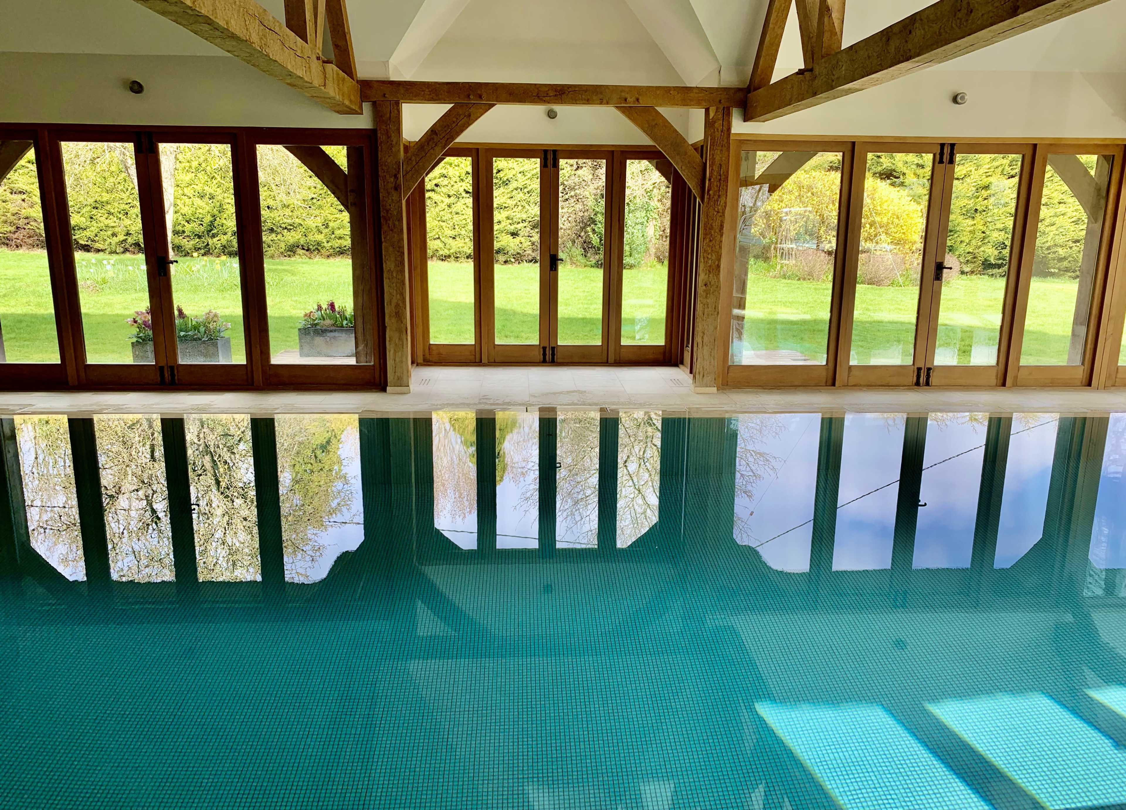 A modern indoor pool area with wooden beams and large glass doors leading to a grassy outdoor space.