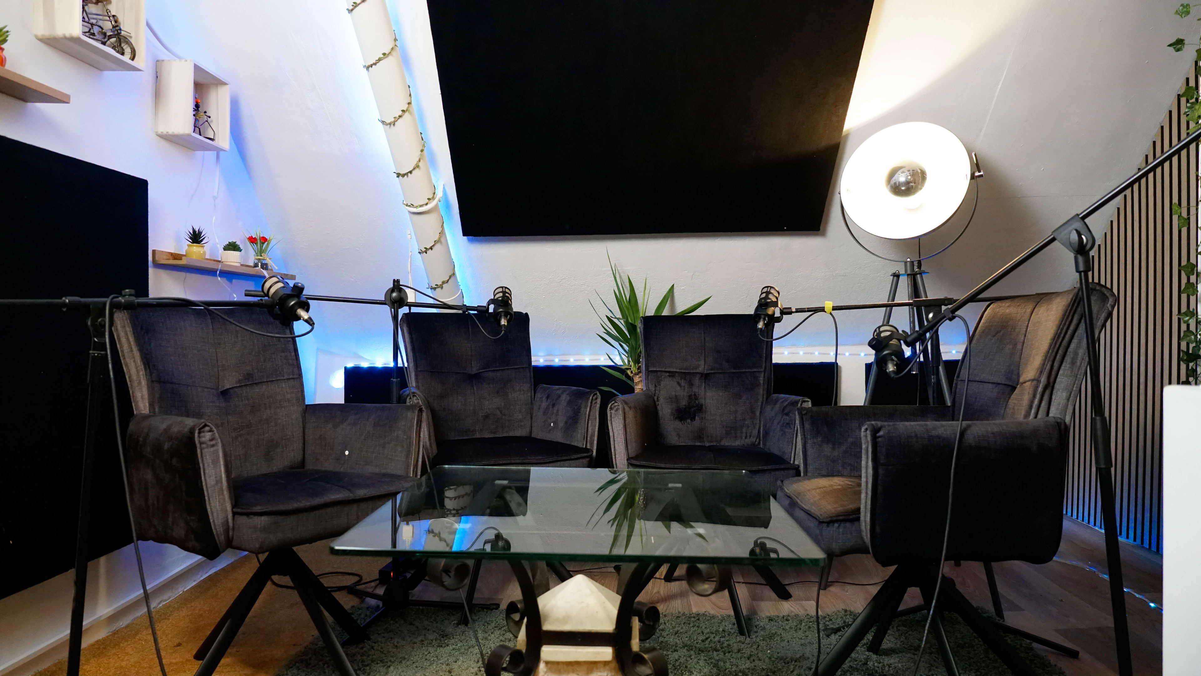 The image shows a cozy podcast recording studio featuring four chairs arranged around a glass table, with microphones set up on stands and ambient lighting in the background.