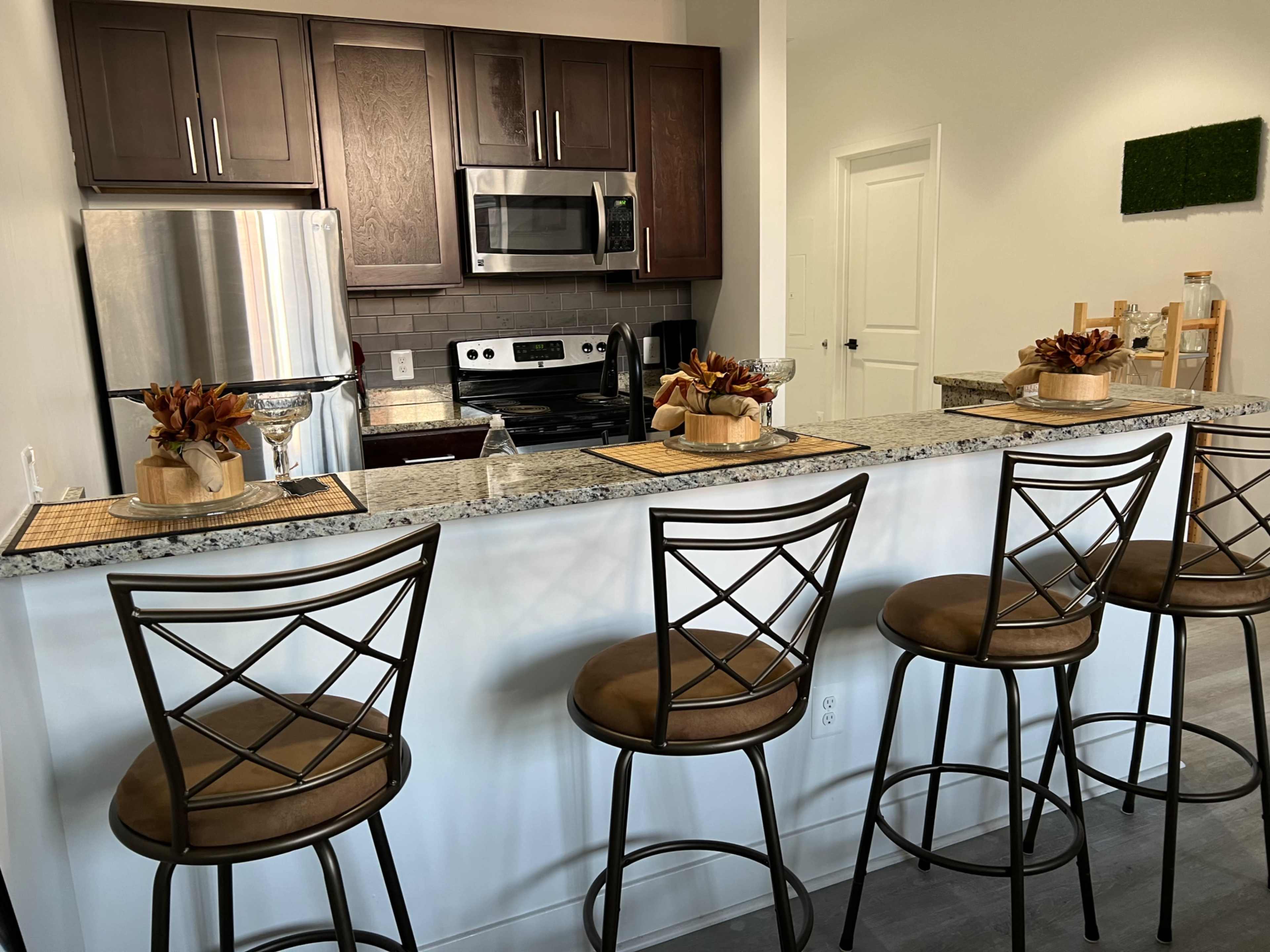 A modern kitchen features a bar with four high stools, granite countertops, and stainless steel appliances.