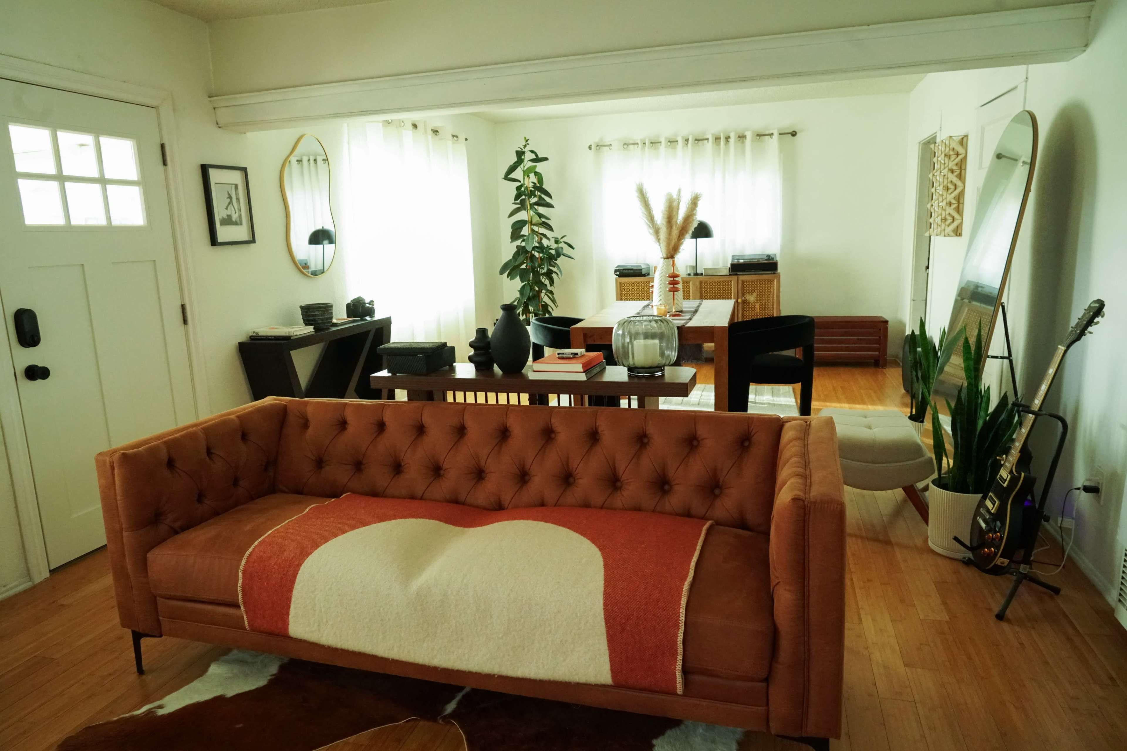 The image features a well-lit living room with a brown tufted sofa, a dining table in the background, and various decorative plants.