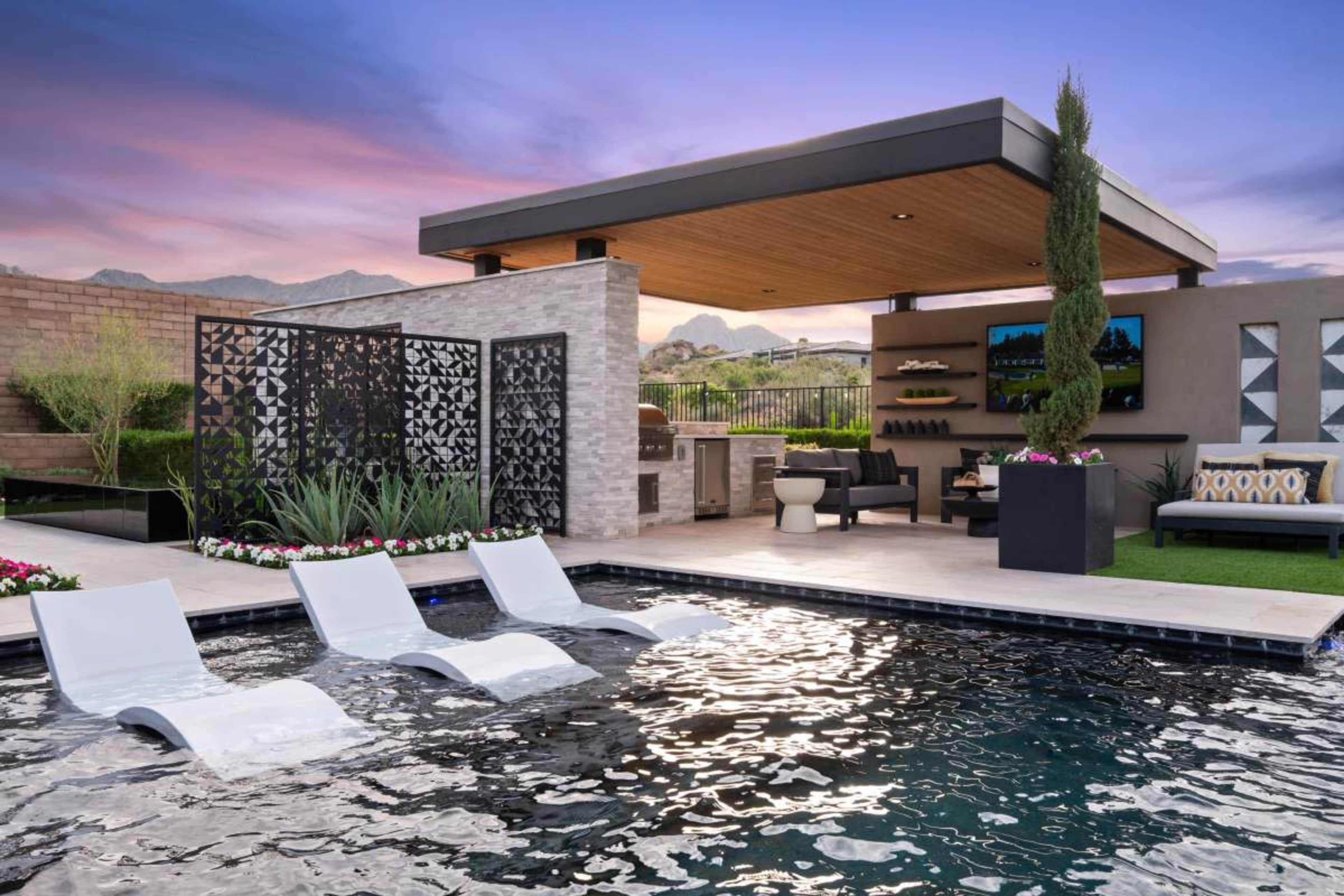 The image shows a modern outdoor pool area featuring lounge chairs partially submerged in the water, a covered seating area with a television, and landscaped plants.