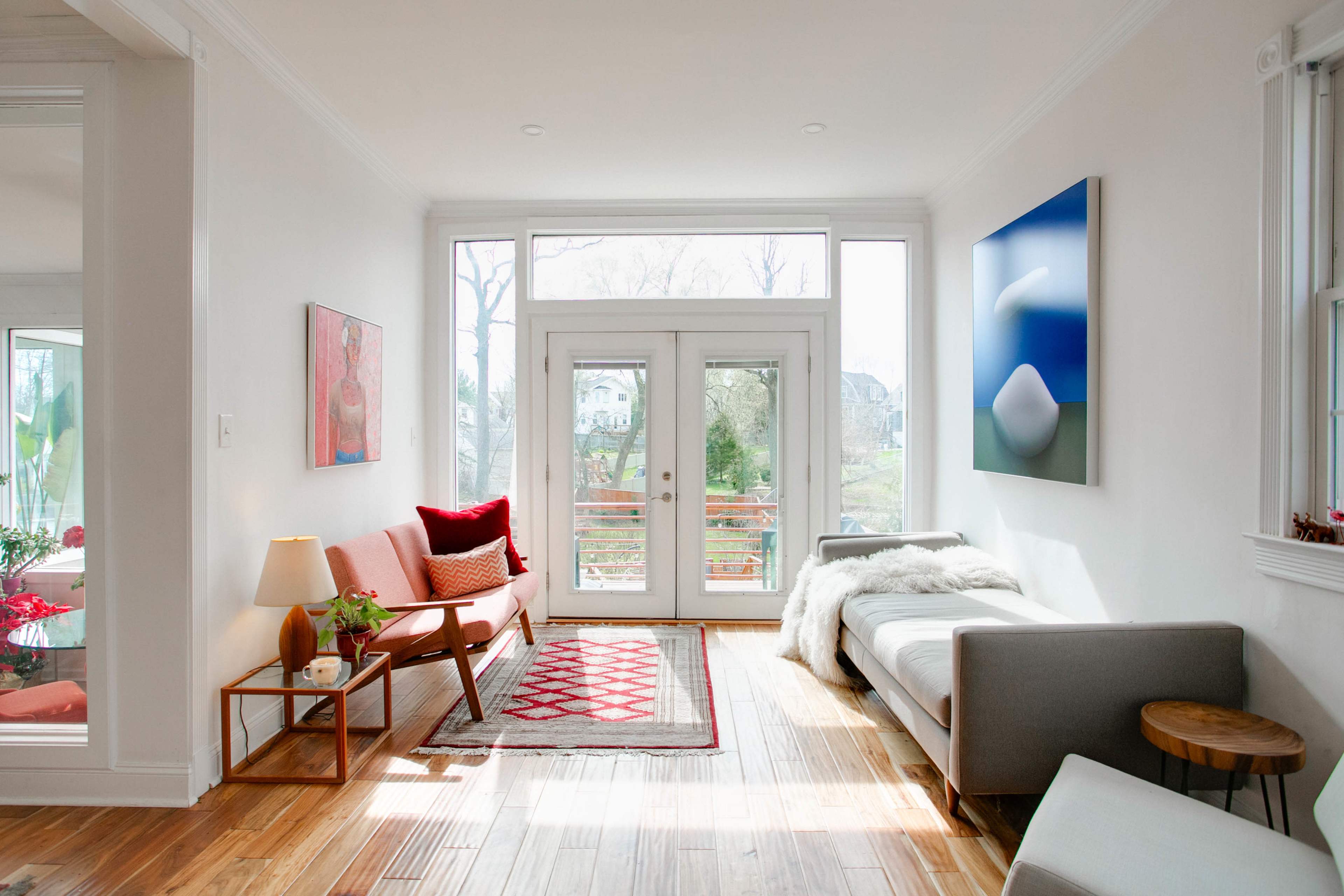 A bright living room features a pink sofa, a gray couch, a coffee table, and large windows overlooking a garden.