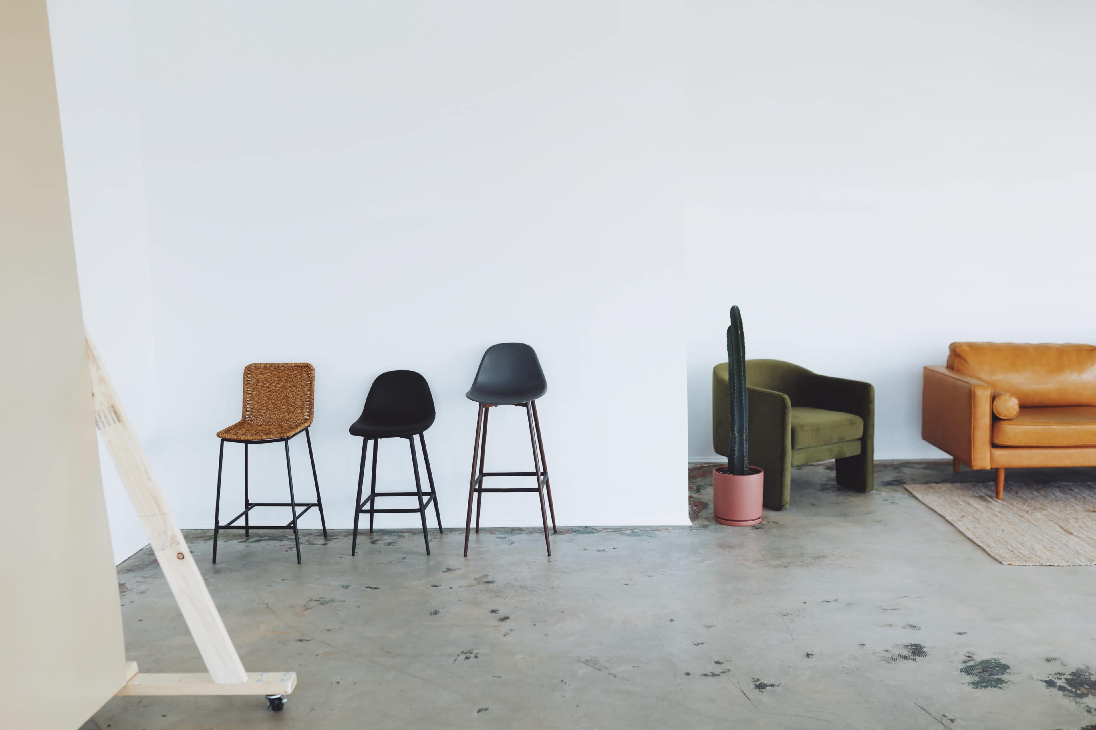 A minimalist interior with three bar stools lined against a white wall, a green armchair, a brown leather sofa, and a potted cactus.