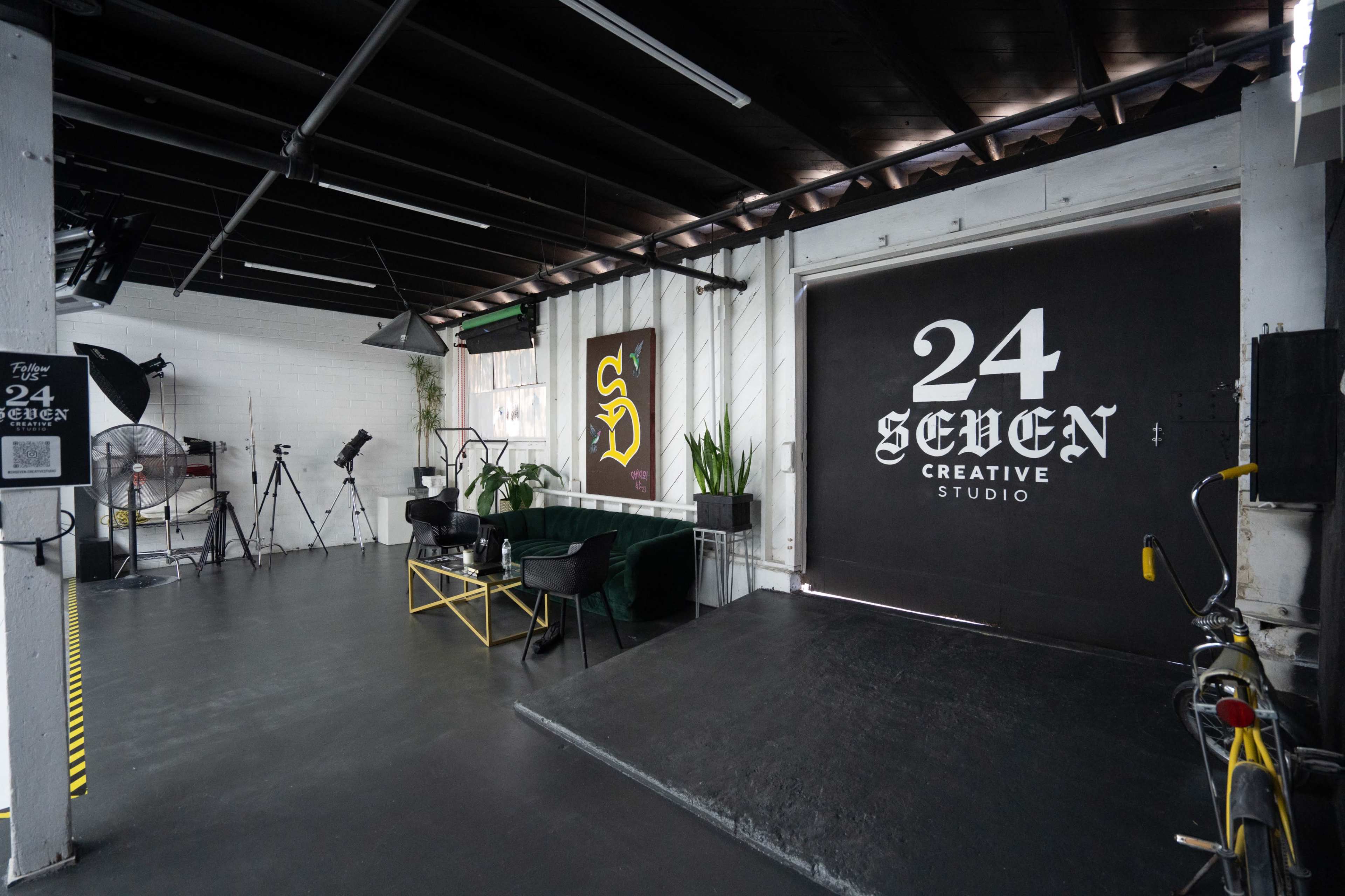 The image shows a creative studio with modern decor, featuring a large black wall with the text "24 SEVEN," green furniture, and various photography equipment arranged around the space.