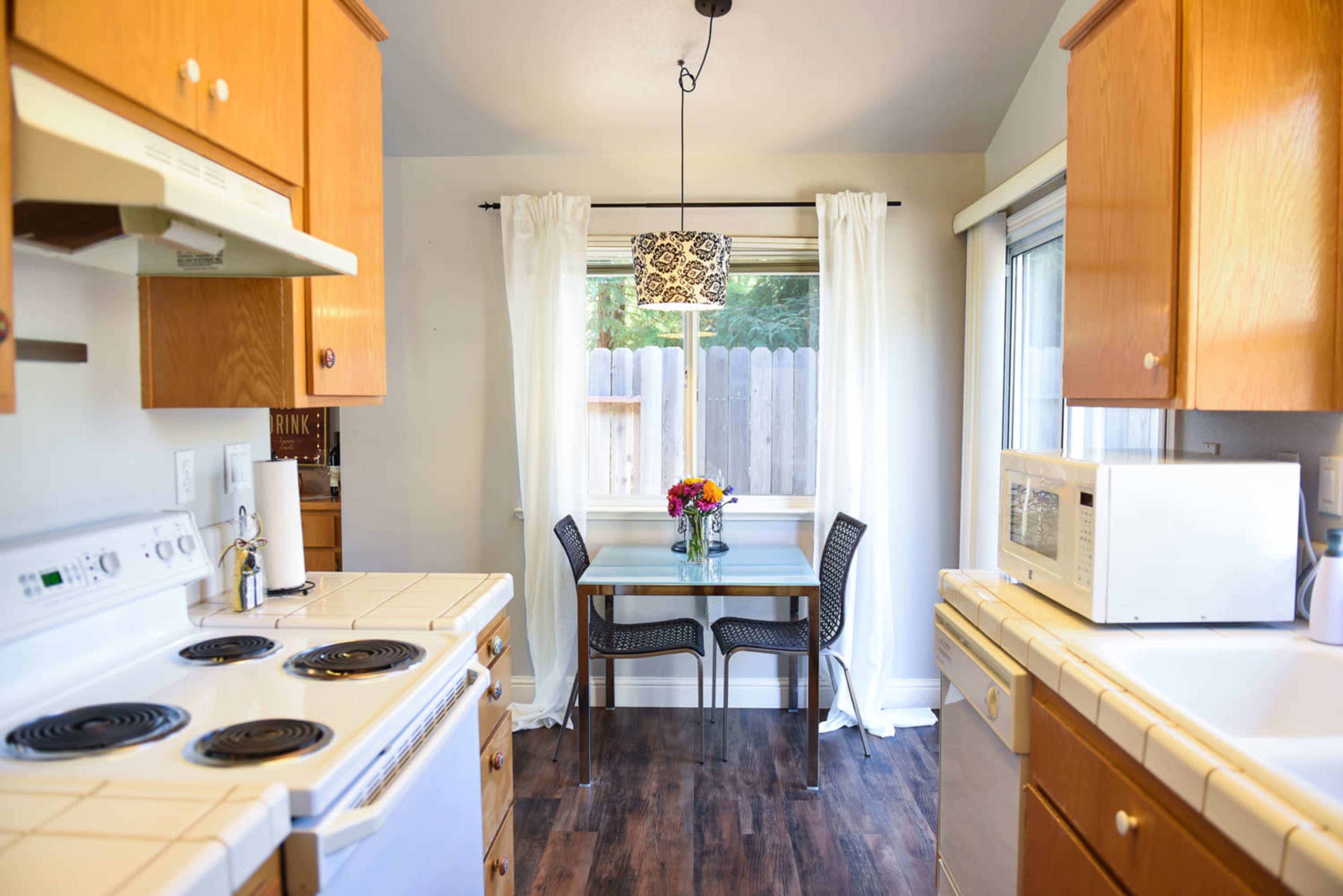 A small kitchen features wooden cabinets, a tiled countertop, and a dining table set for two with a vase of flowers by a window.
