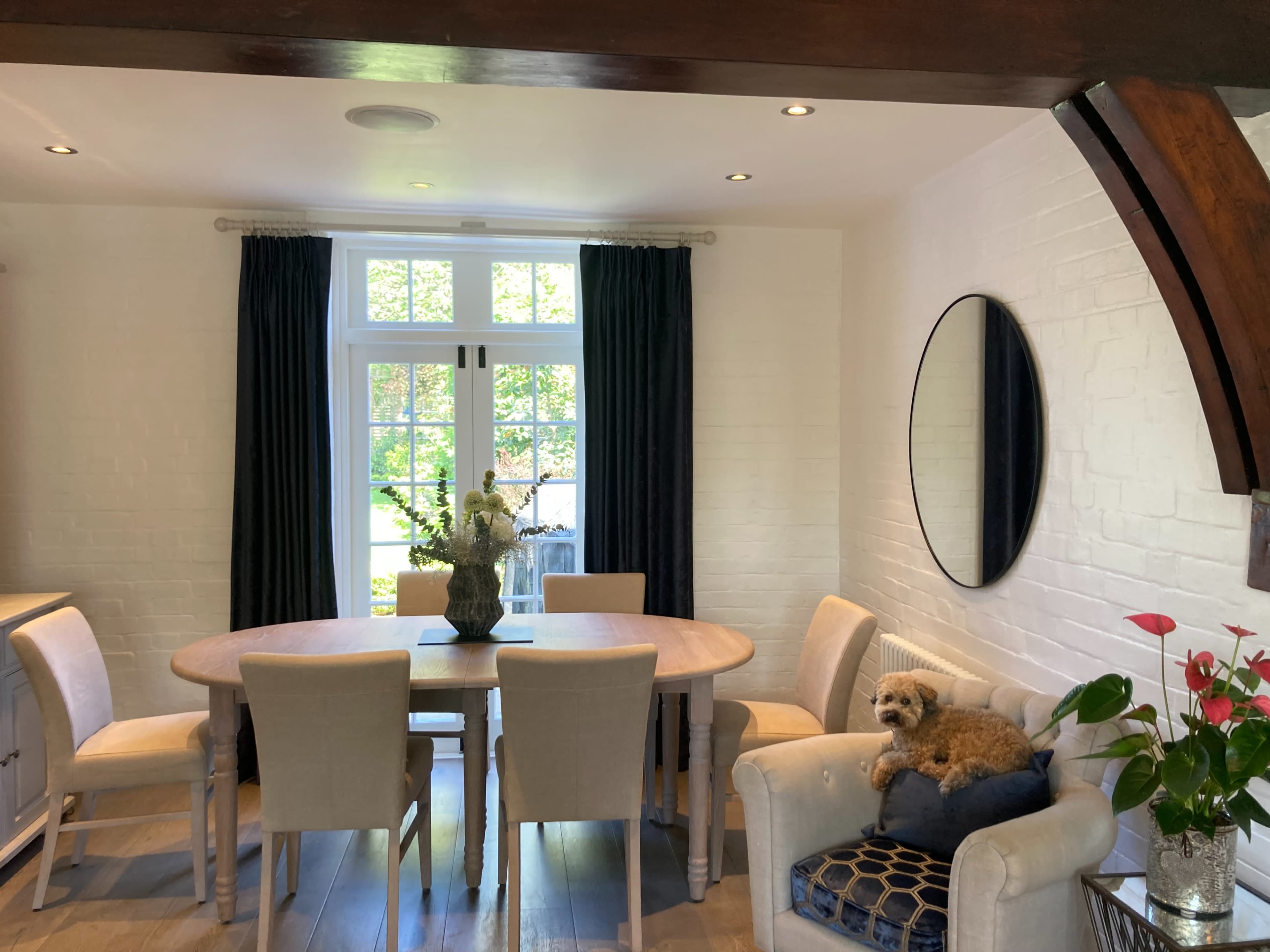 A dining area with a round wooden table surrounded by upholstered chairs, a large mirror on the wall, and a small dog resting on a chair.