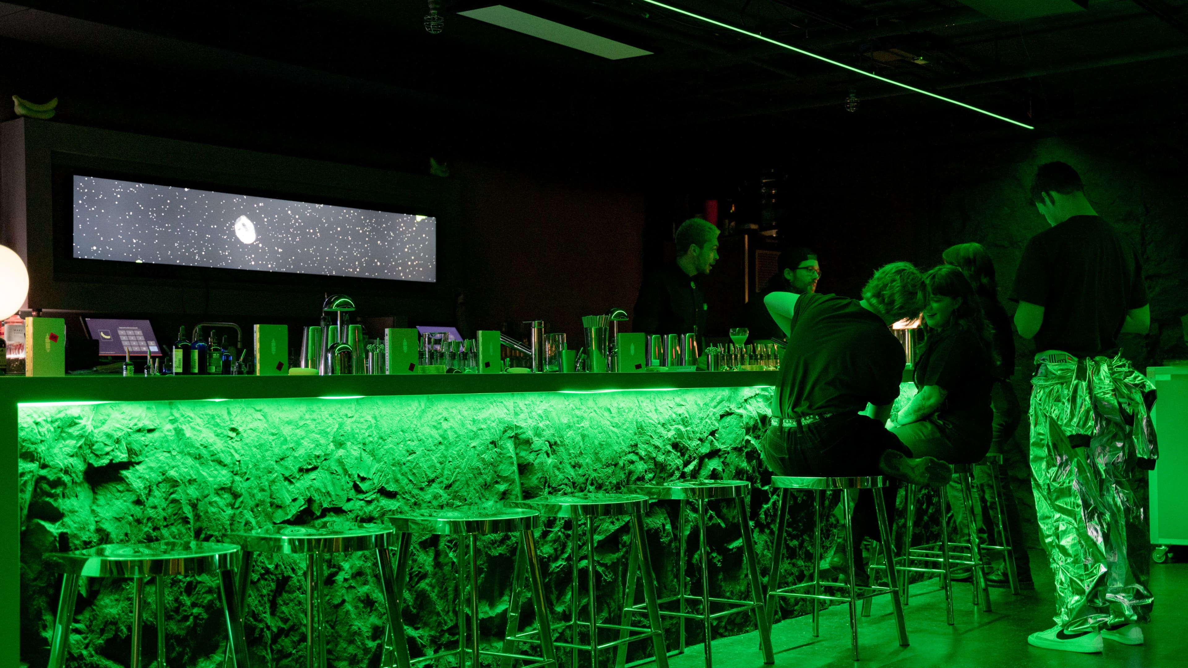 A group of people sits at a bar with a green-lit stone wall, while others stand behind the counter amid a starry screen display.