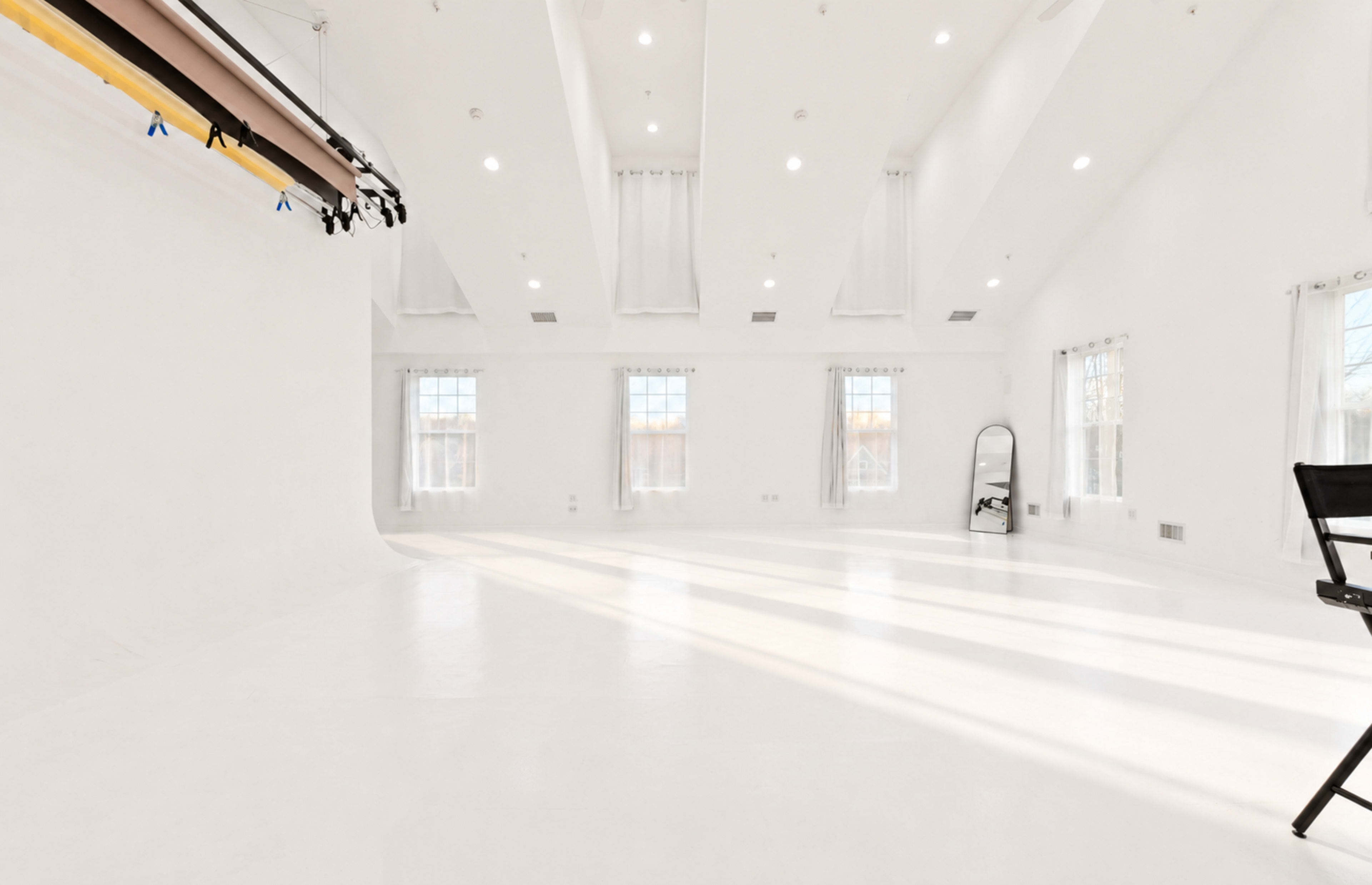 The image shows a spacious, well-lit studio with white walls, a high ceiling, and large windows, featuring a mirror and a director's chair.