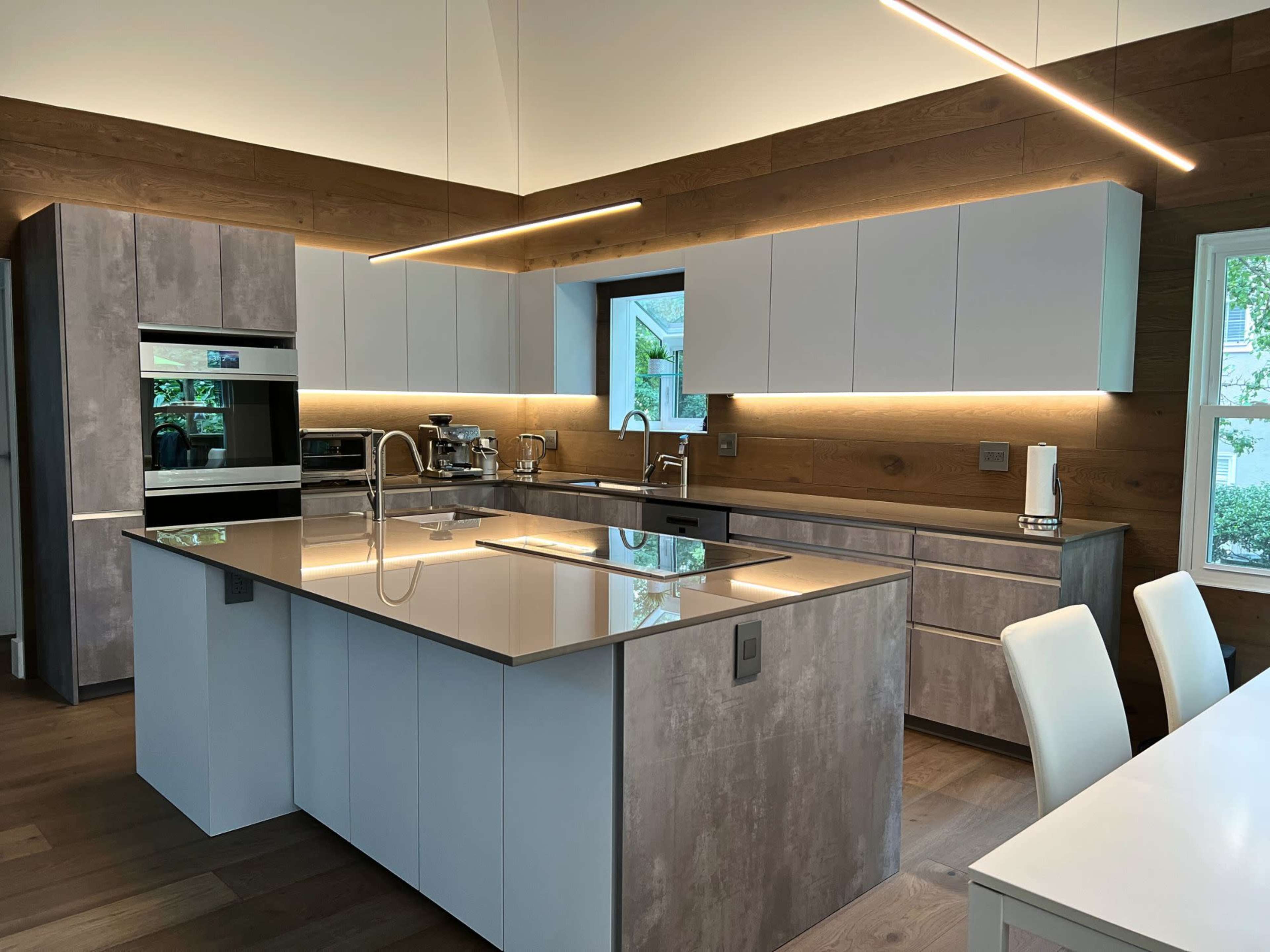 The image shows a modern kitchen with a central island, featuring a combination of light wood and gray cabinetry, under-cabinet lighting, and stainless steel appliances.