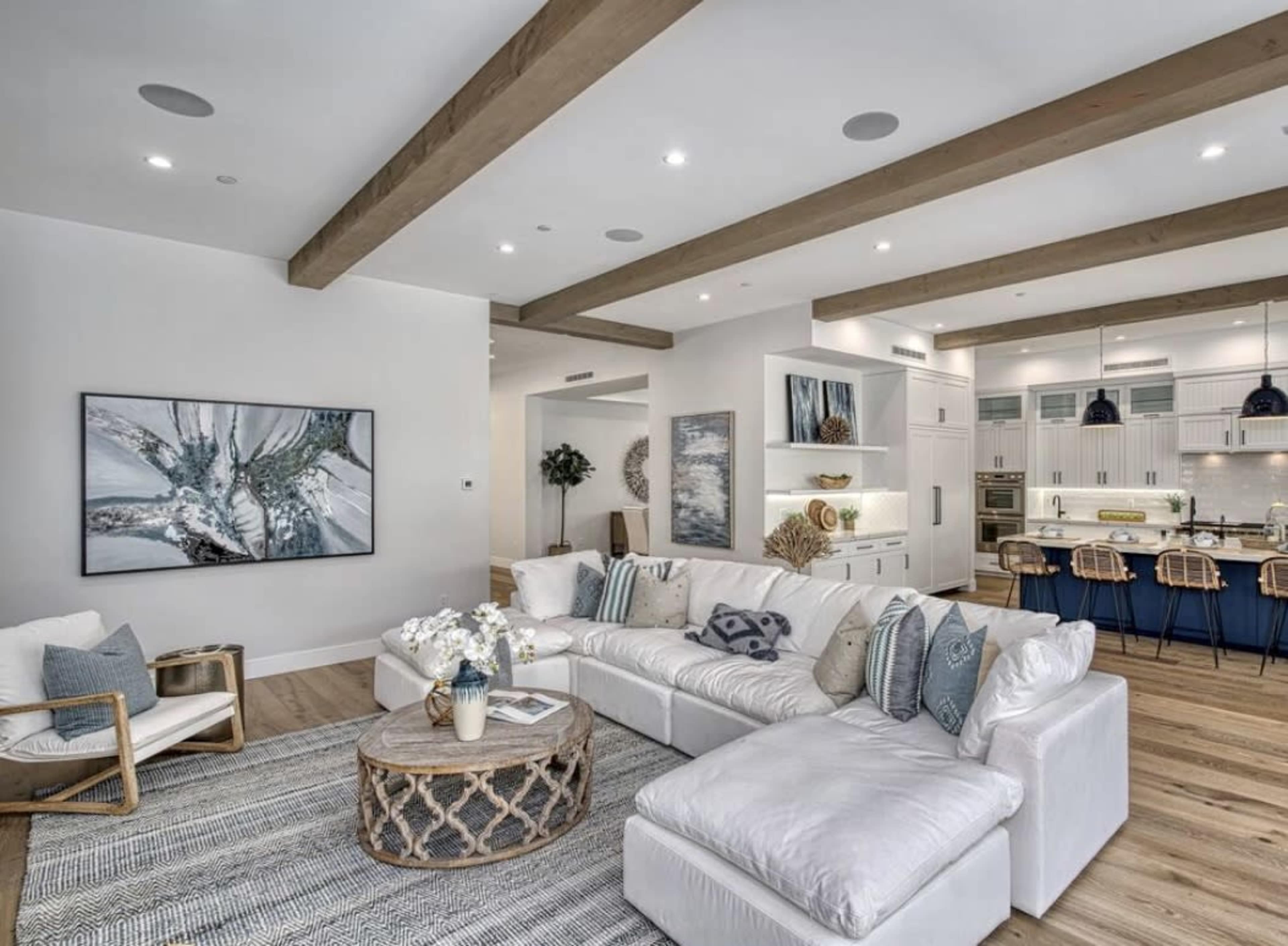 A spacious living room with a large sectional sofa, a round coffee table, and a kitchen area in the background featuring white cabinetry and dark bar stools.