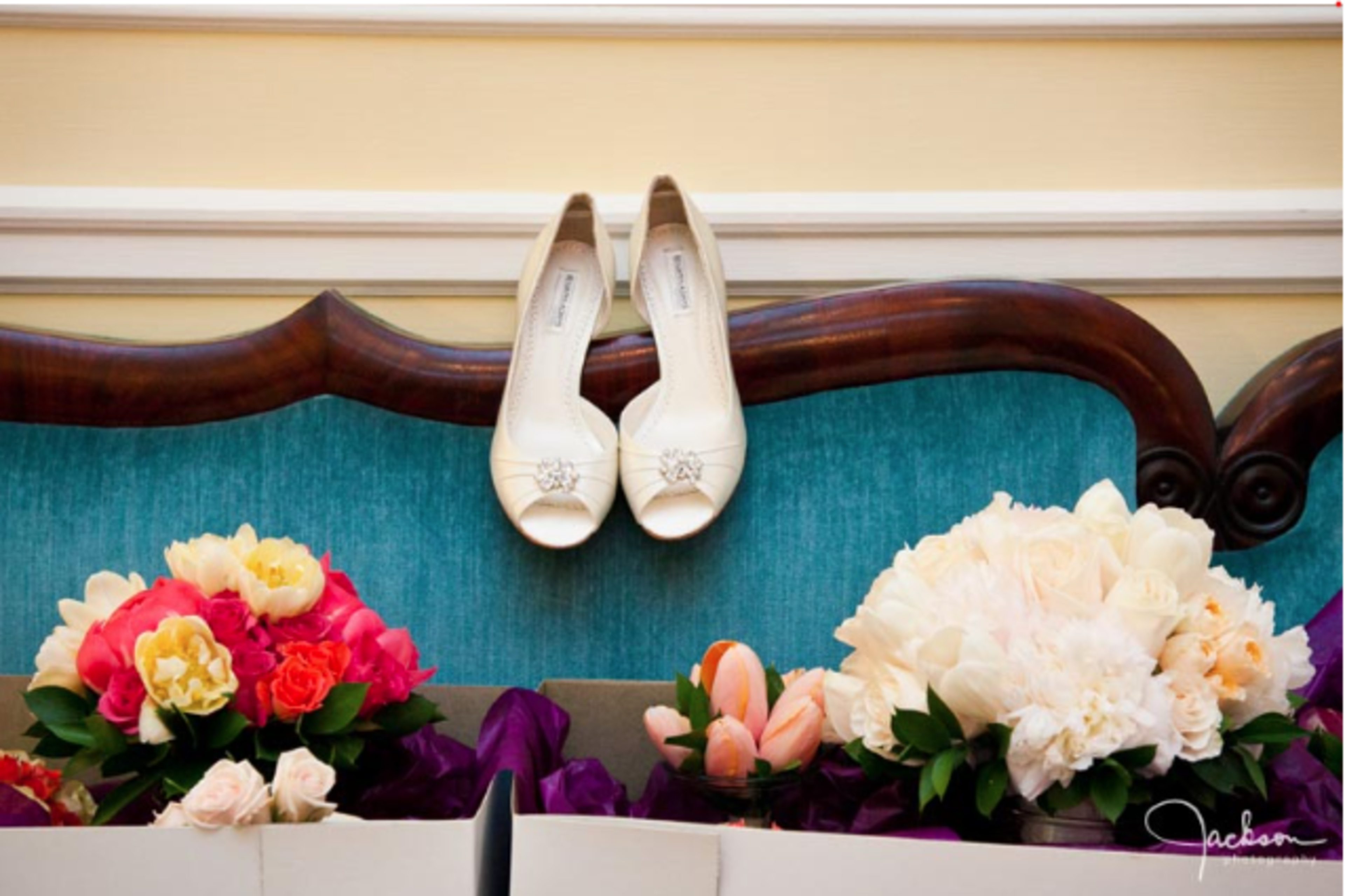 A pair of white high-heeled shoes is placed above a collection of vibrant floral arrangements on a decorative backdrop.