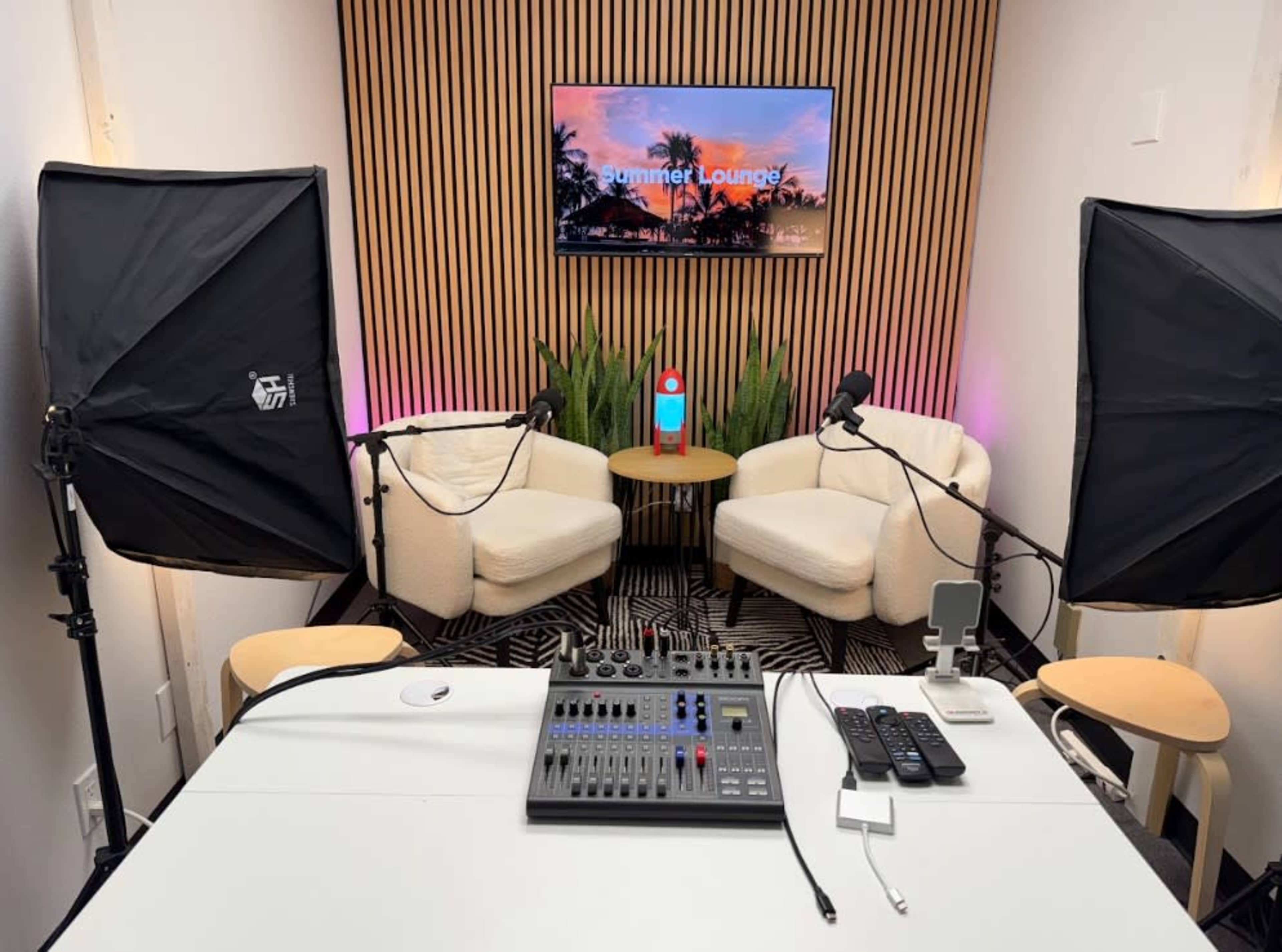 A well-equipped podcast studio features two cushioned chairs facing a table with audio equipment, flanked by large softbox lights and a television displaying a tropical scene.