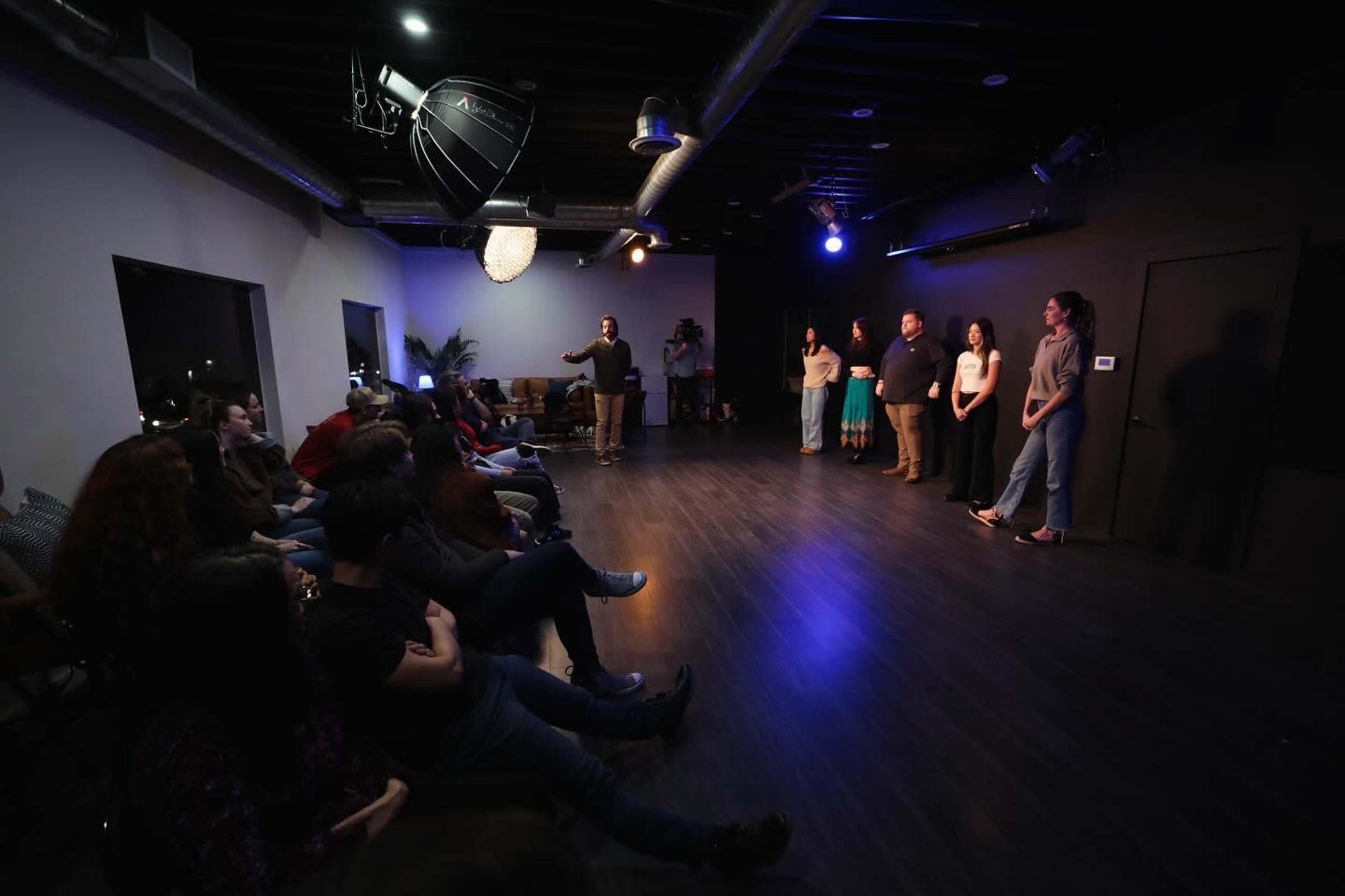 A group of performers stands on stage while an audience watches from seated positions in a dimly lit room.