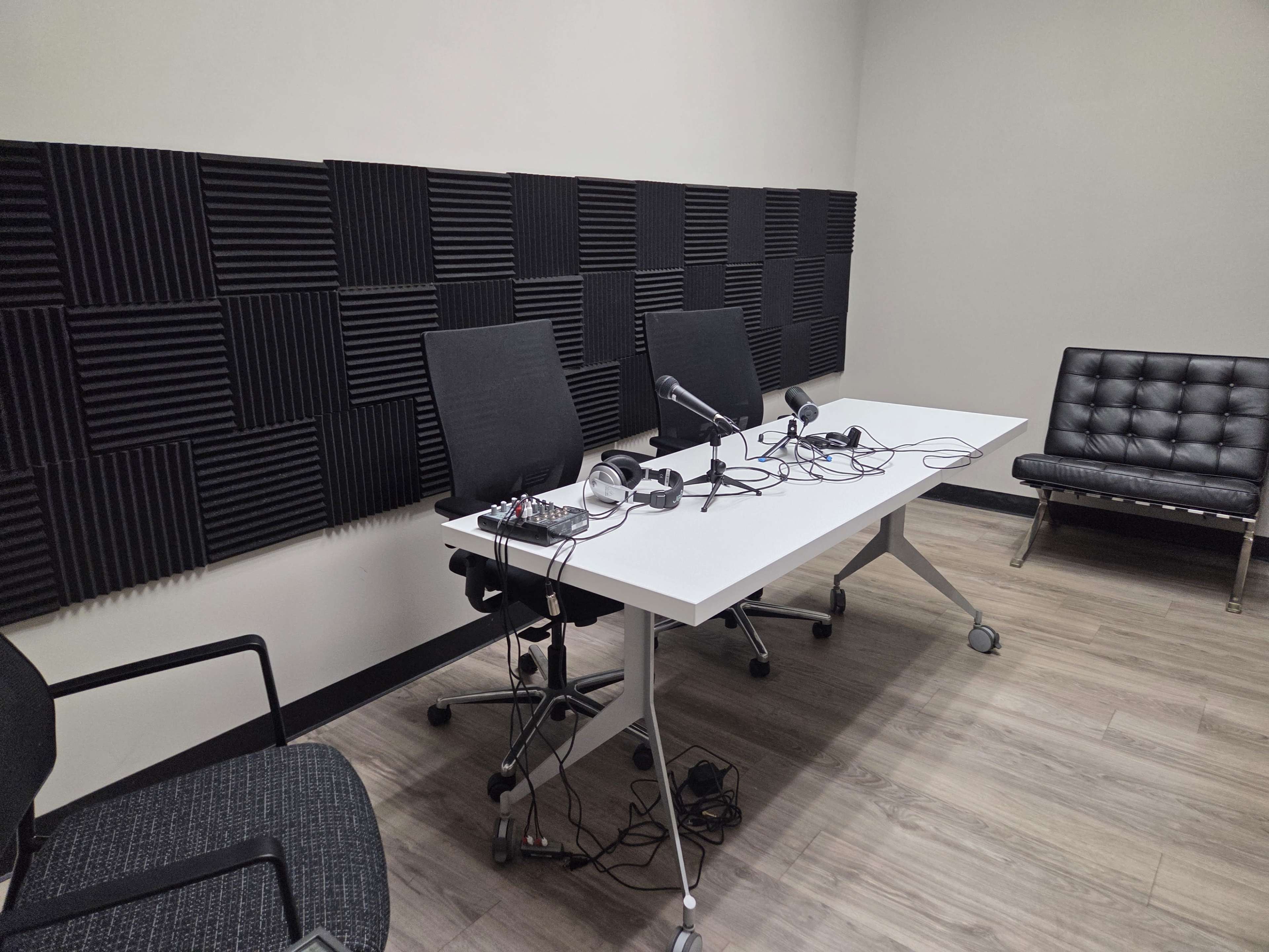 The image shows a small recording studio with a white table, two chairs, microphones, and soundproofing panels on the walls.