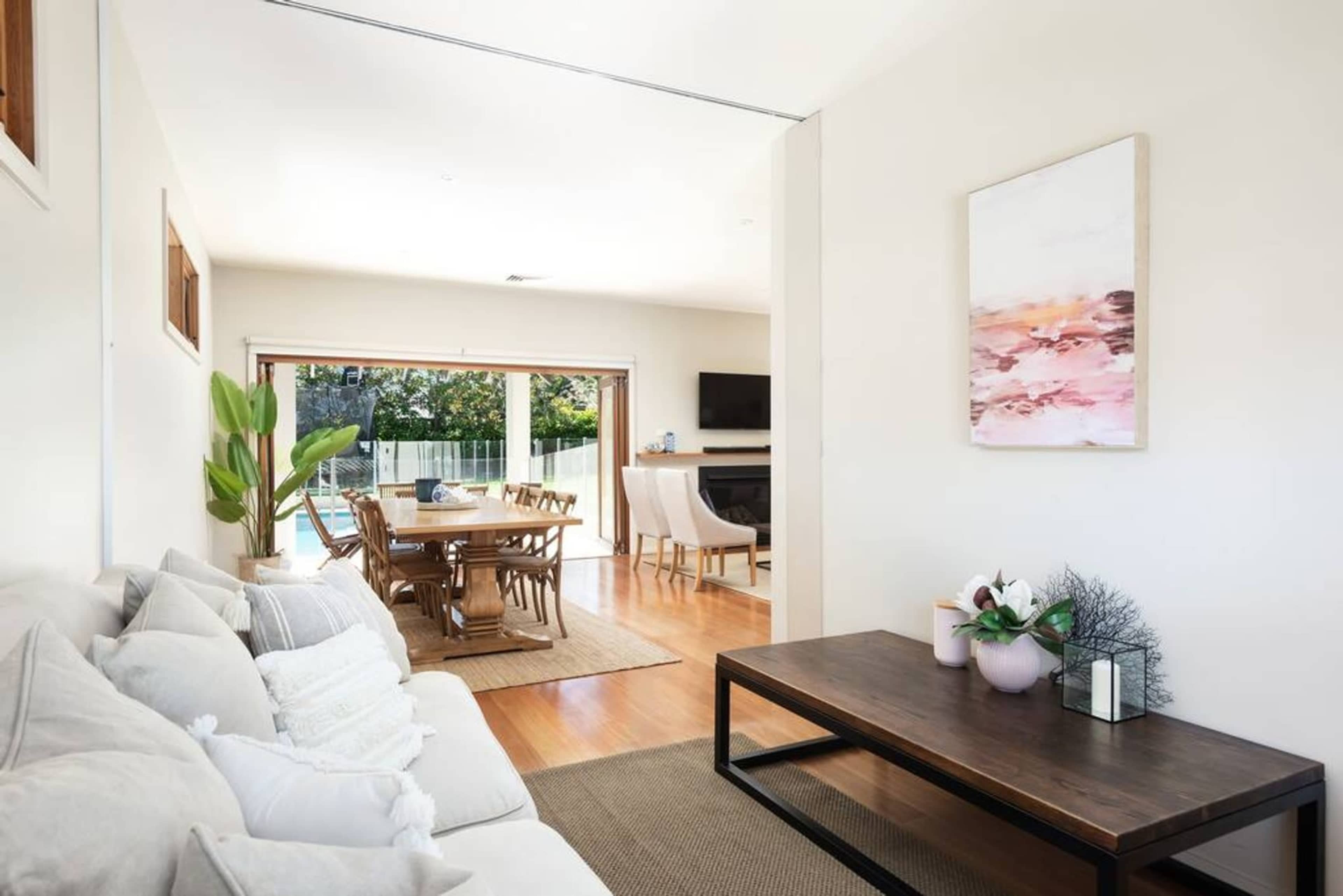 A bright, open living area with a sofa, coffee table, and a dining space that leads to a patio with a pool in the background.