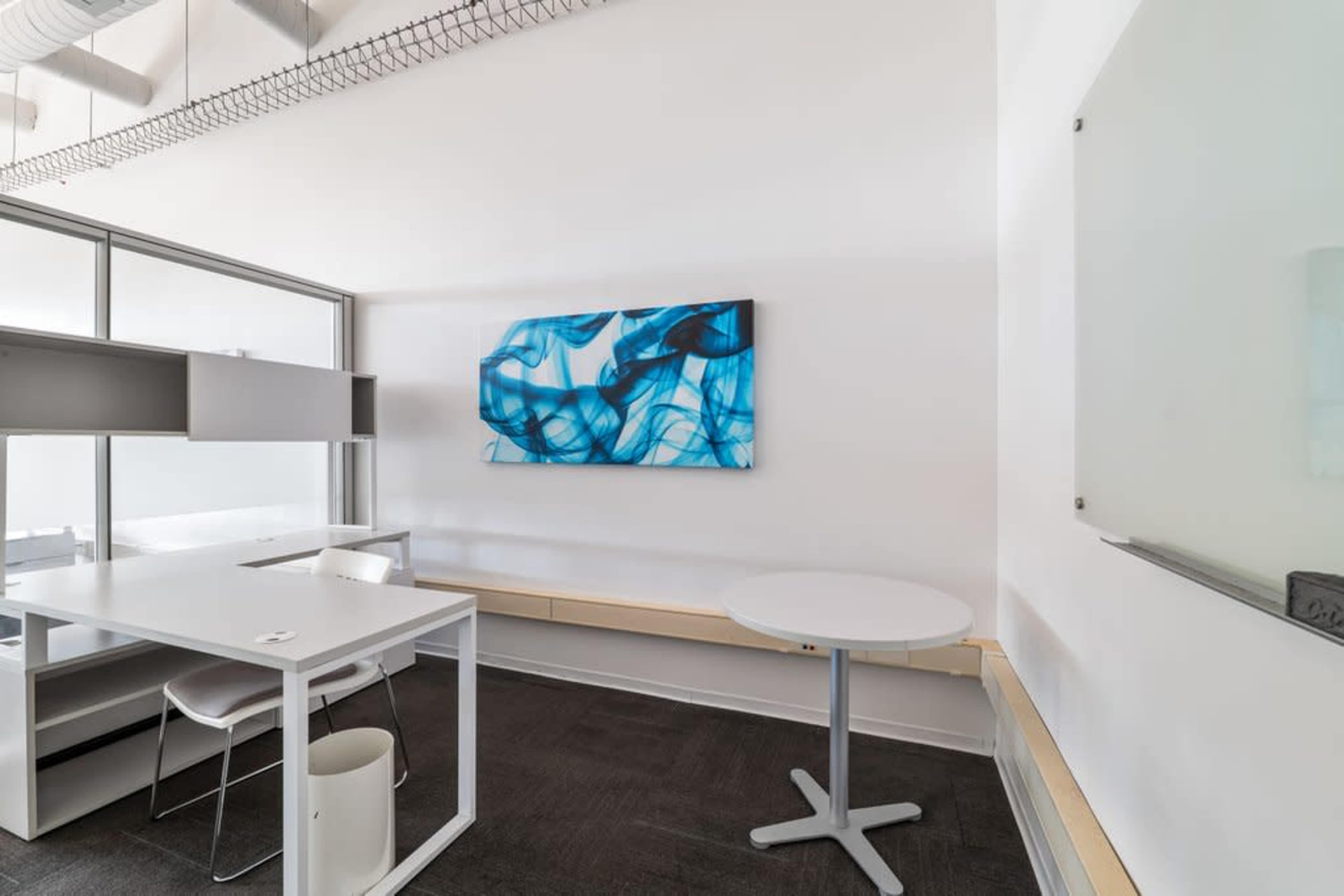 A minimalist office space features a white table, a round table, and a blue abstract artwork on the wall.