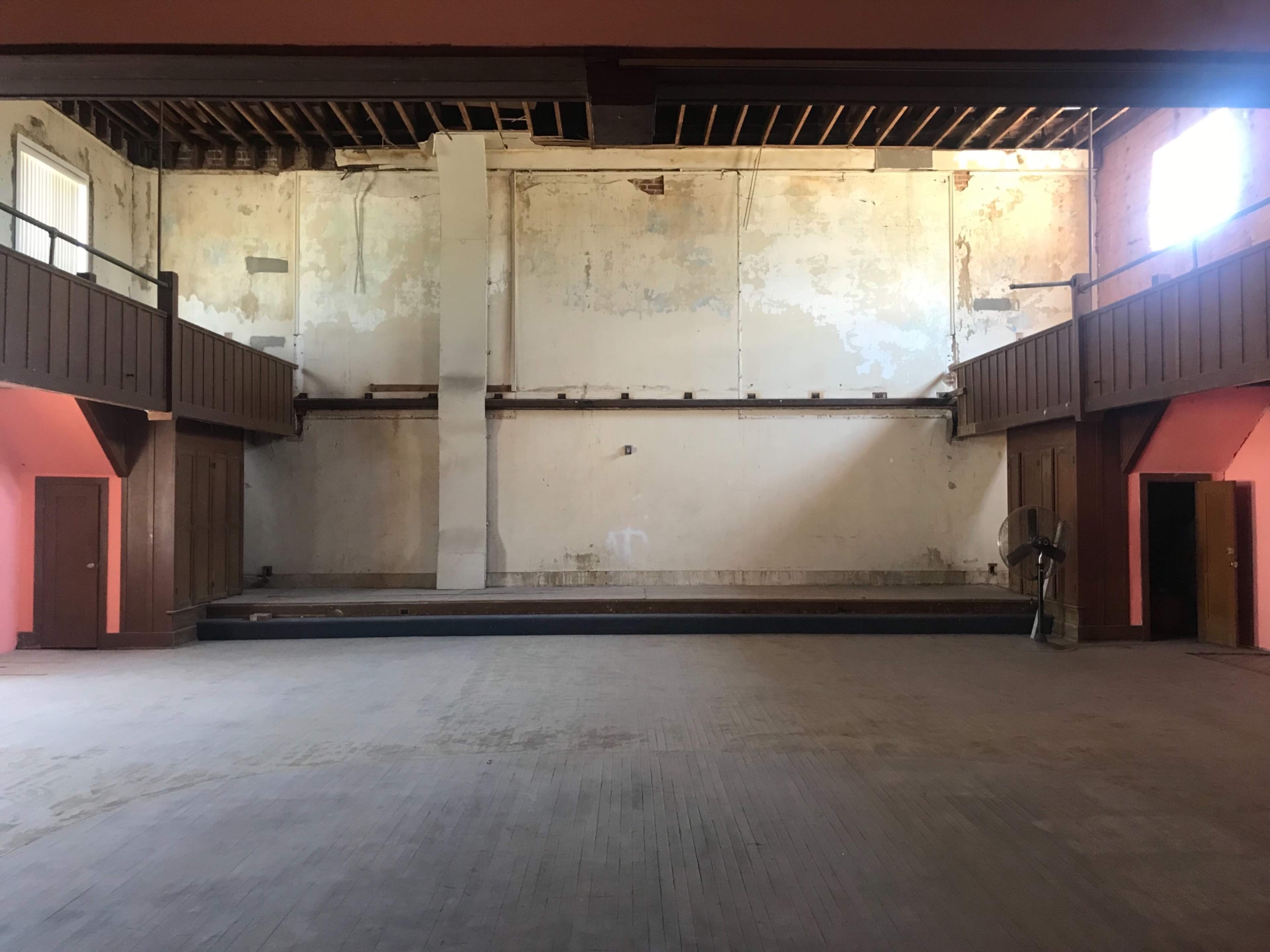 An empty, dilapidated interior of a large room with peeling walls, wooden balconies on either side, and a stage at the far end.