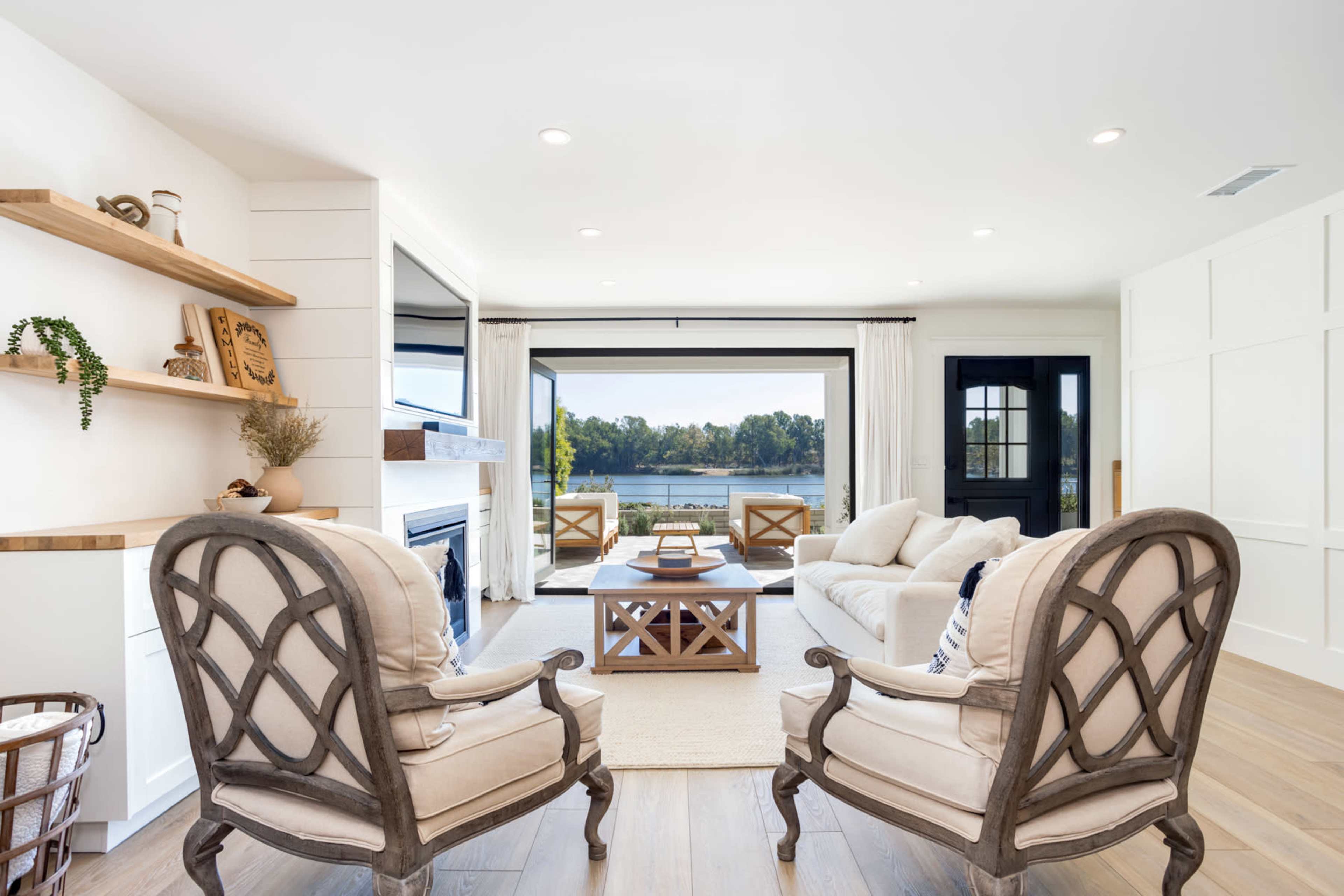 A modern living room with a light color scheme, featuring two patterned armchairs, a coffee table, and large windows overlooking a body of water.