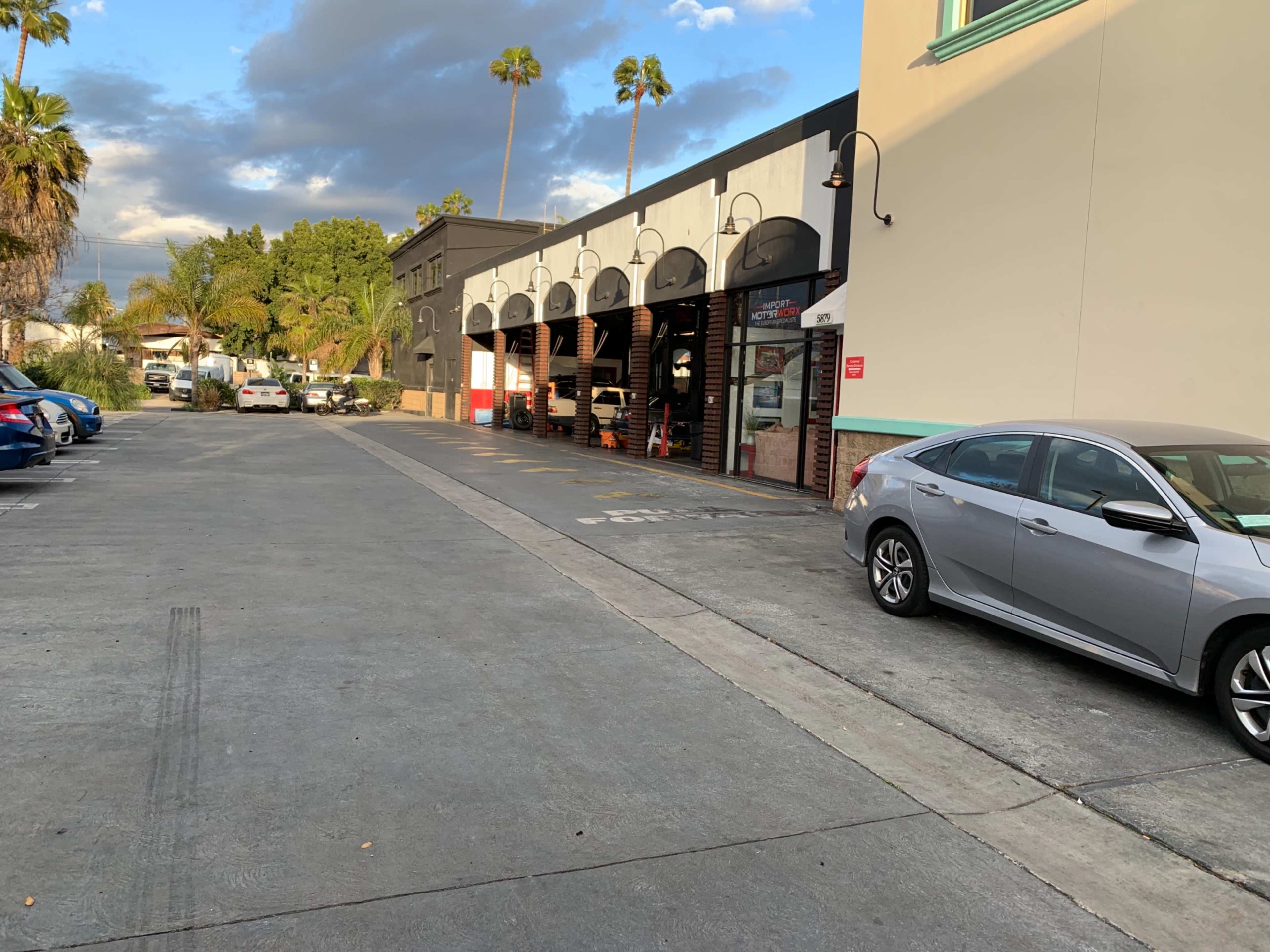 A row of buildings with arched openings lines a paved street, accompanied by parked cars and palm trees in the background.