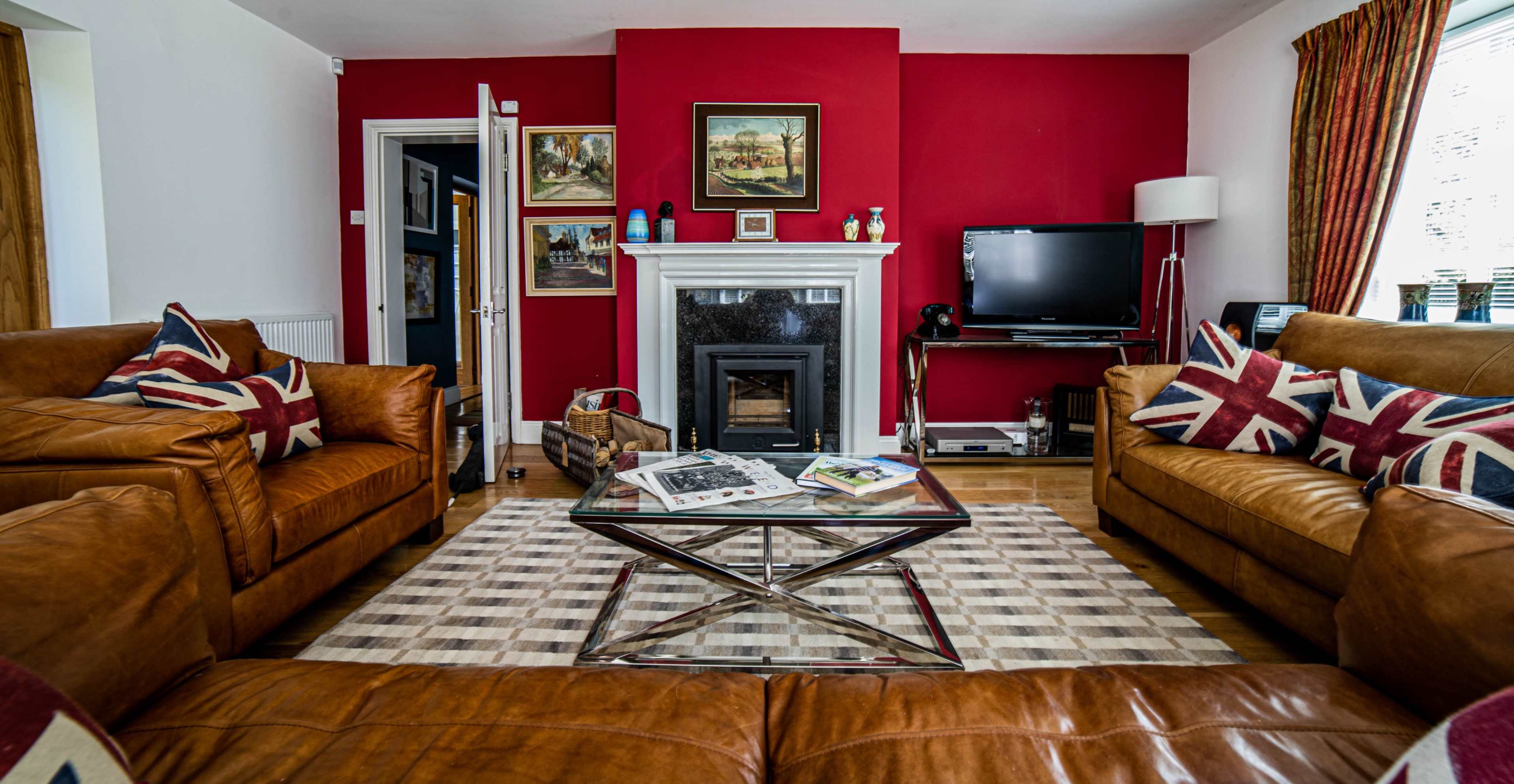 The living room features two brown leather sofas with Union Jack pillows facing a glass coffee table, set against a red wall and a fireplace, with a television and artwork on display.