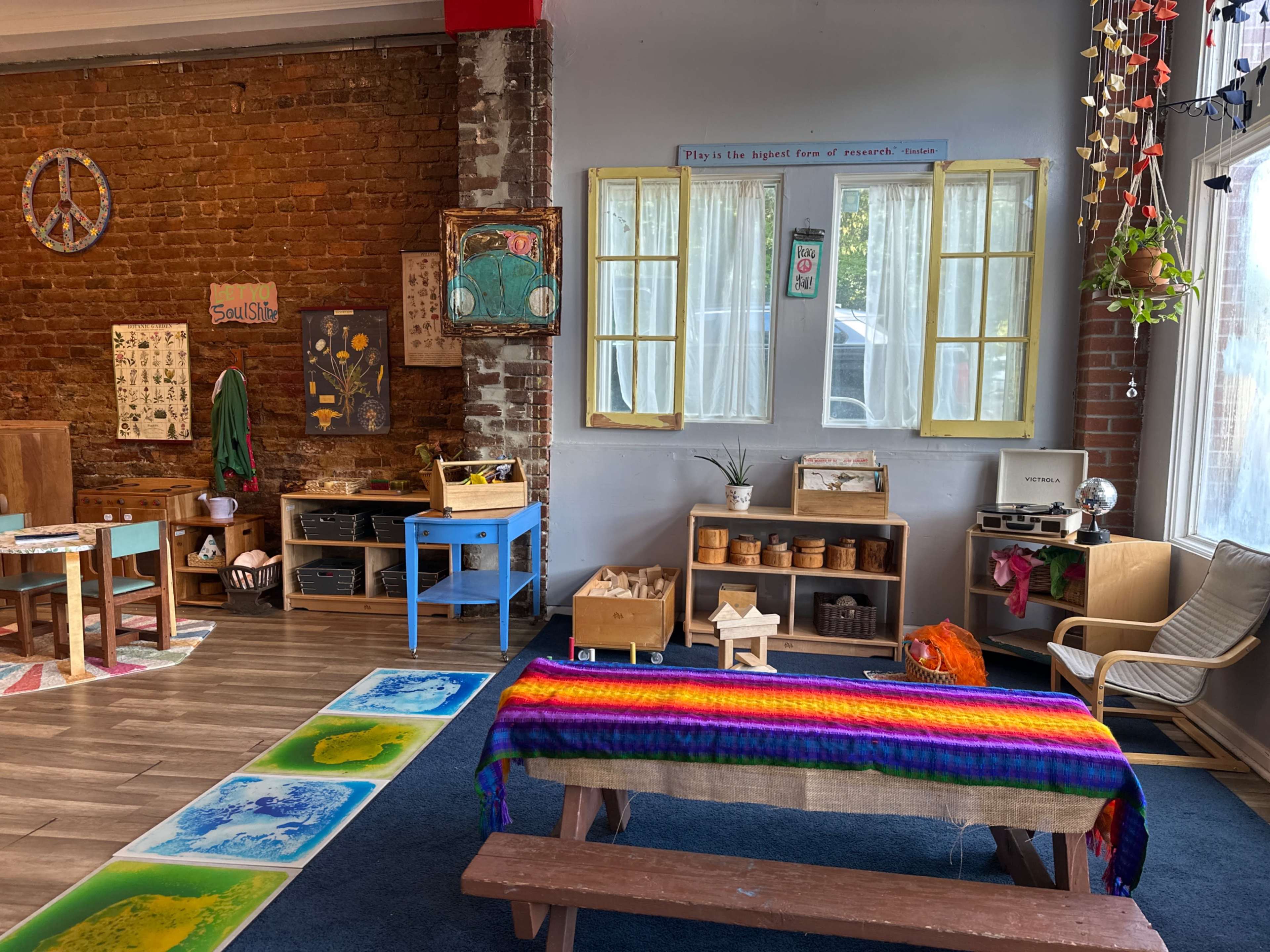 The image shows a colorful, inviting classroom space with wooden furniture, educational materials, and a large window allowing natural light to enter.