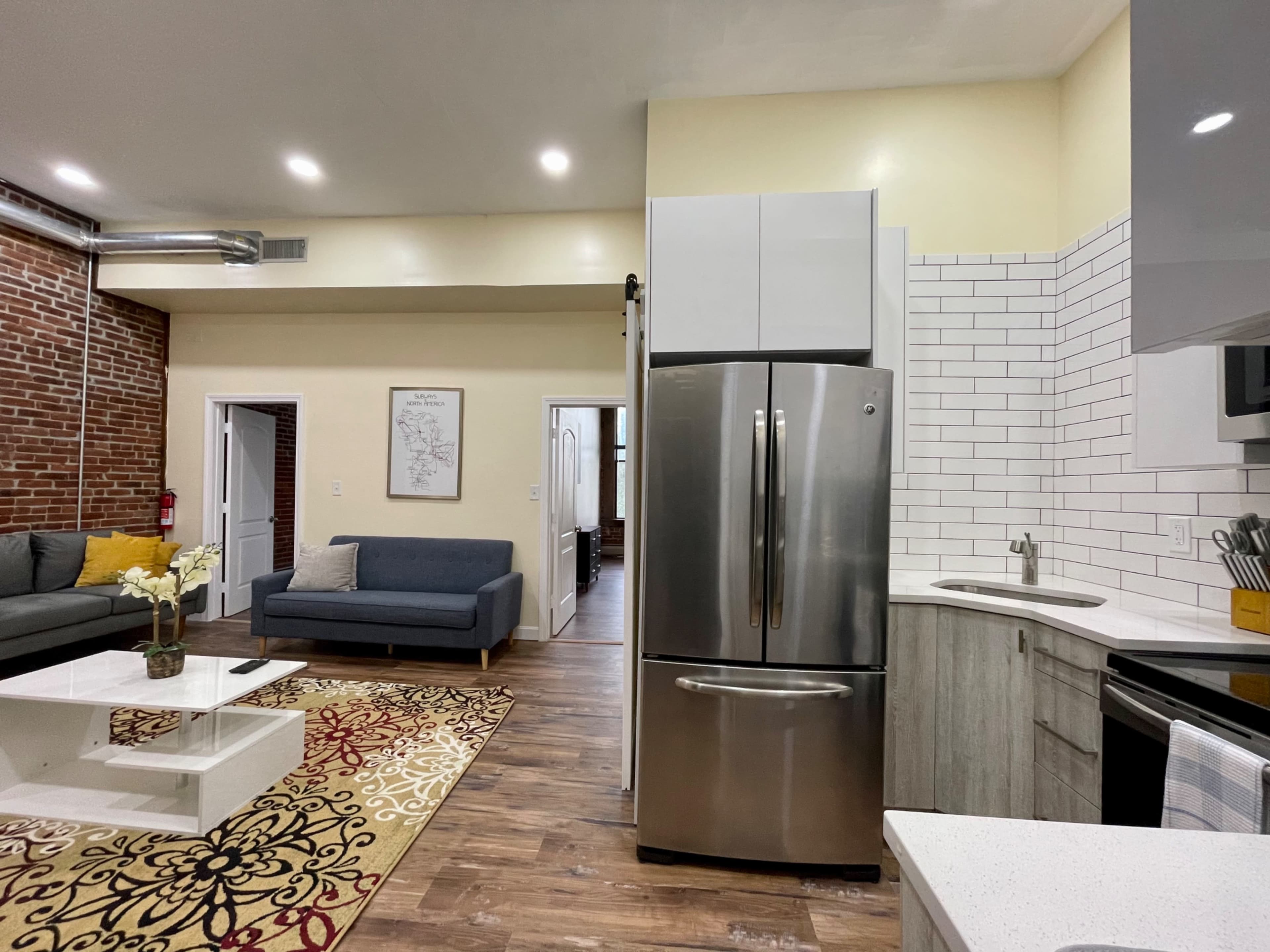 A modern kitchen and living area showcase a stainless steel refrigerator, a brick wall, and contemporary furniture.