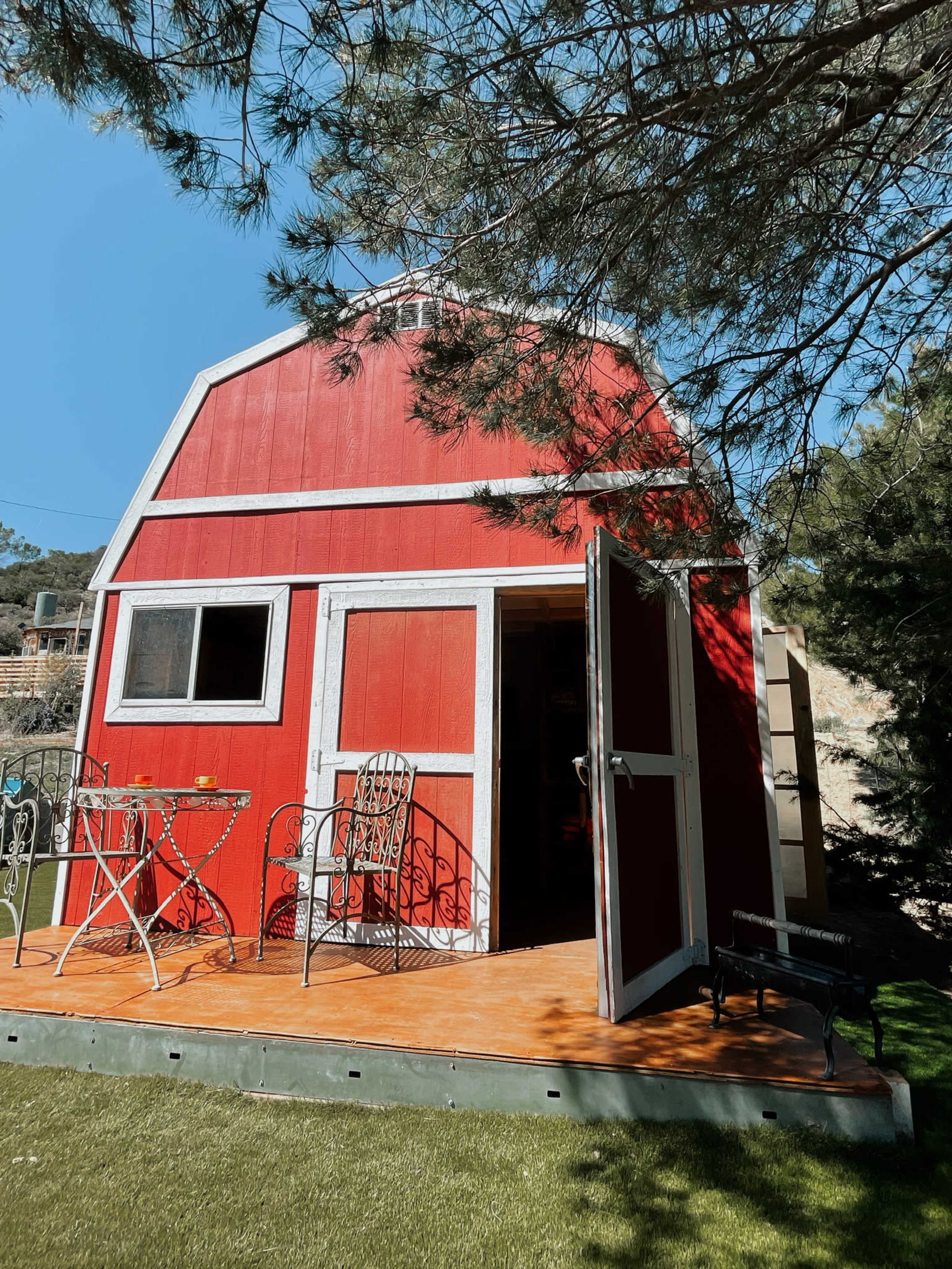 A red barn-shaped structure with an open door and a small table set outside sits among trees.