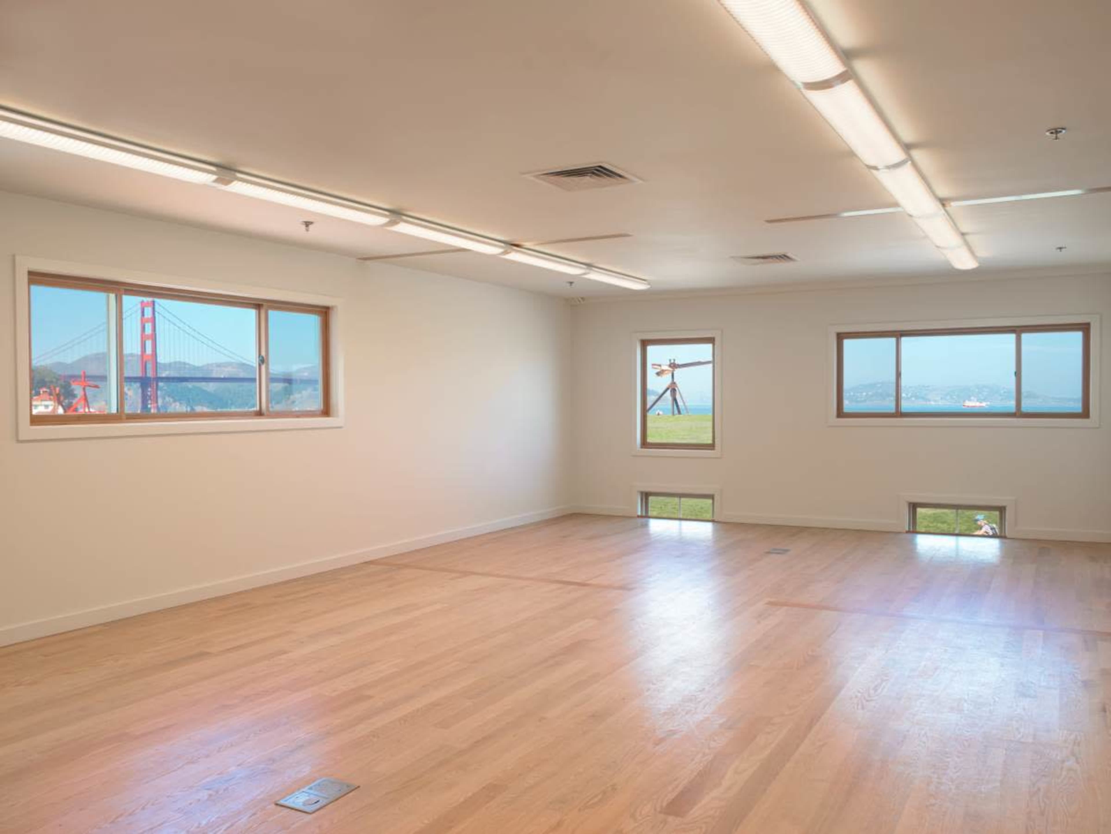 The image shows an empty room with hardwood floors and large windows offering a view of the Golden Gate Bridge.