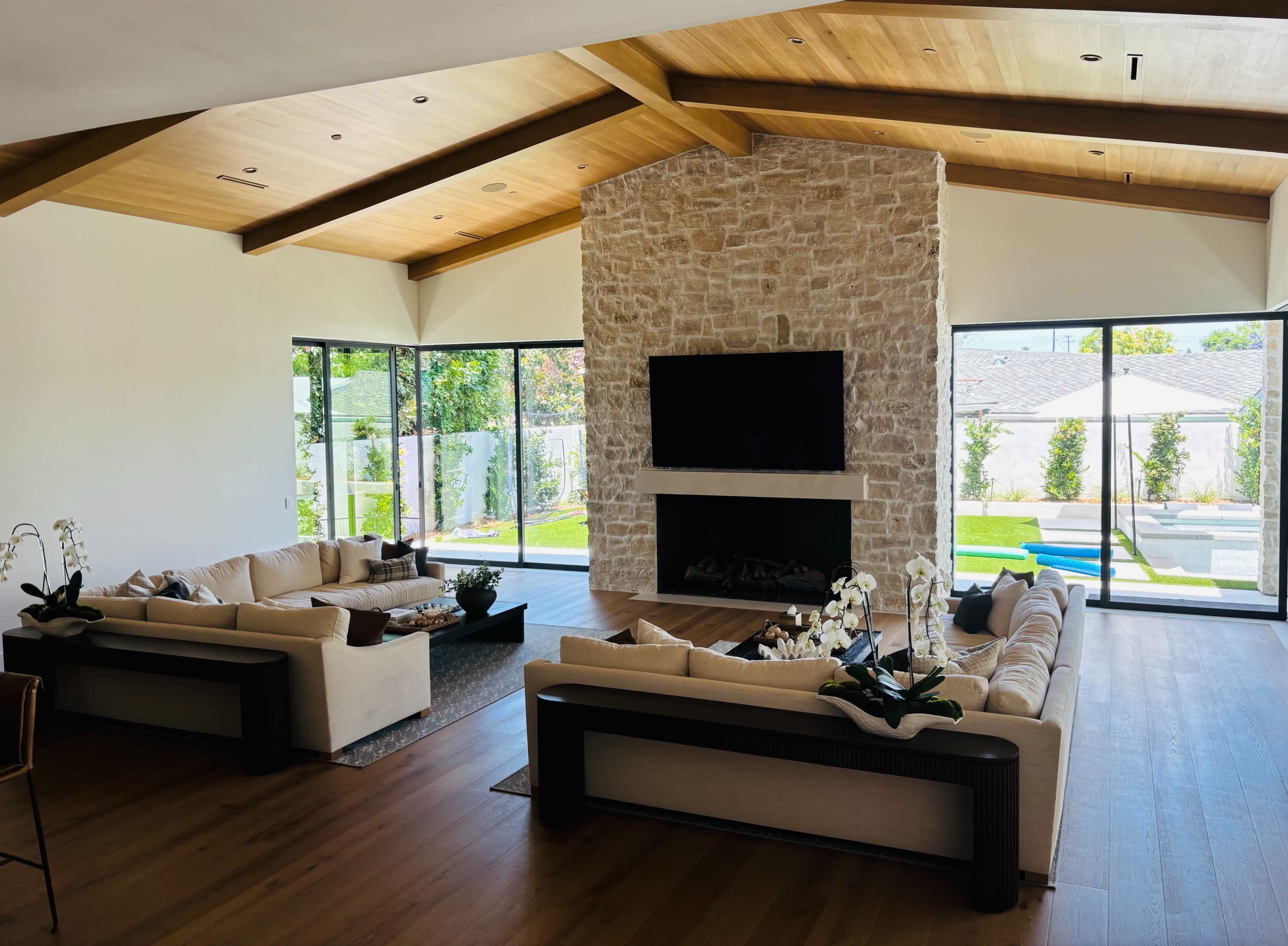 A spacious living room features two large sofas facing a stone fireplace and a wall-mounted television, with large windows providing views of a landscaped backyard.