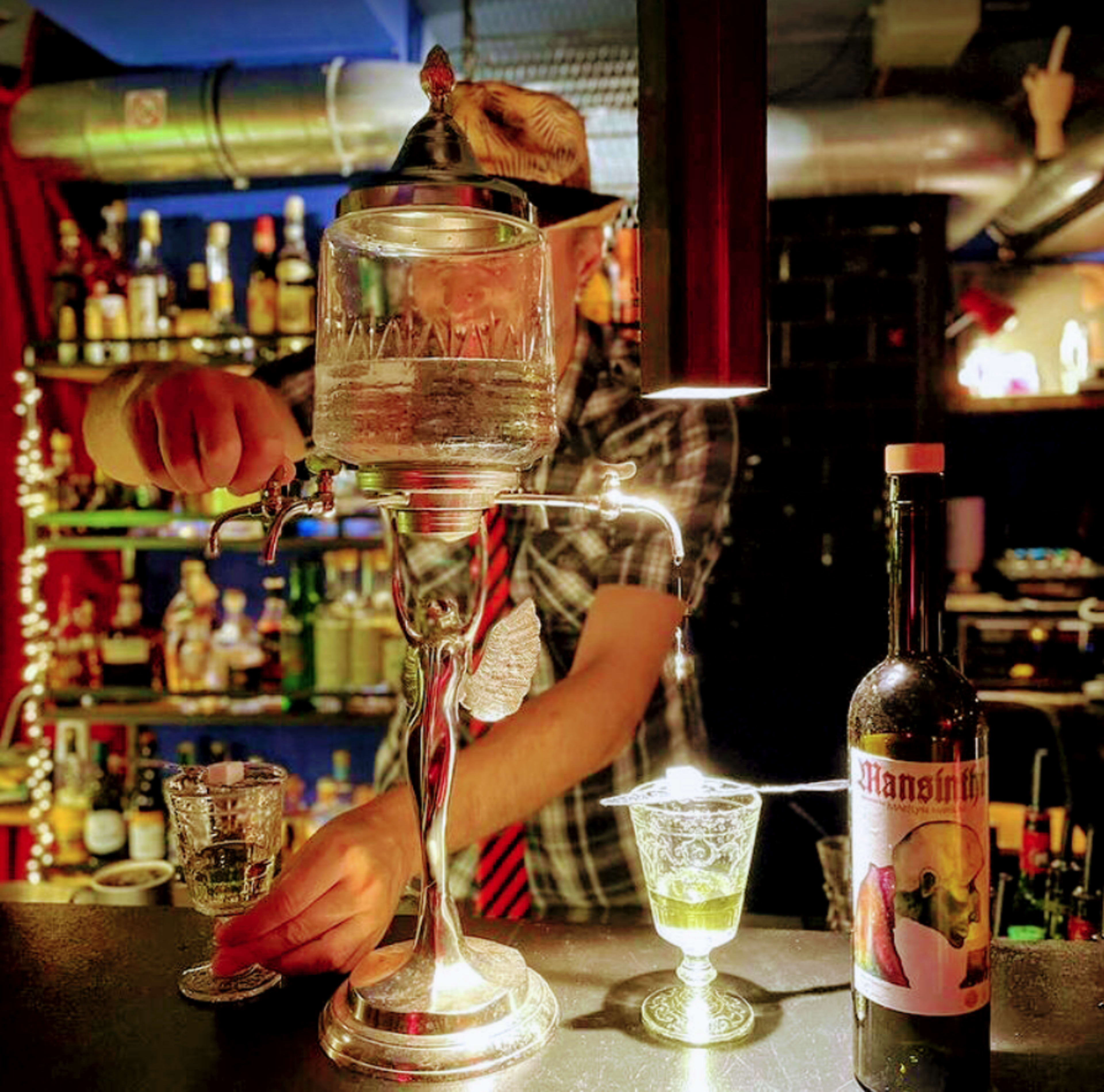 A bartender pours a clear liquid from a vintage dispenser into a glass at a dimly lit bar, with shelves of bottles in the background.