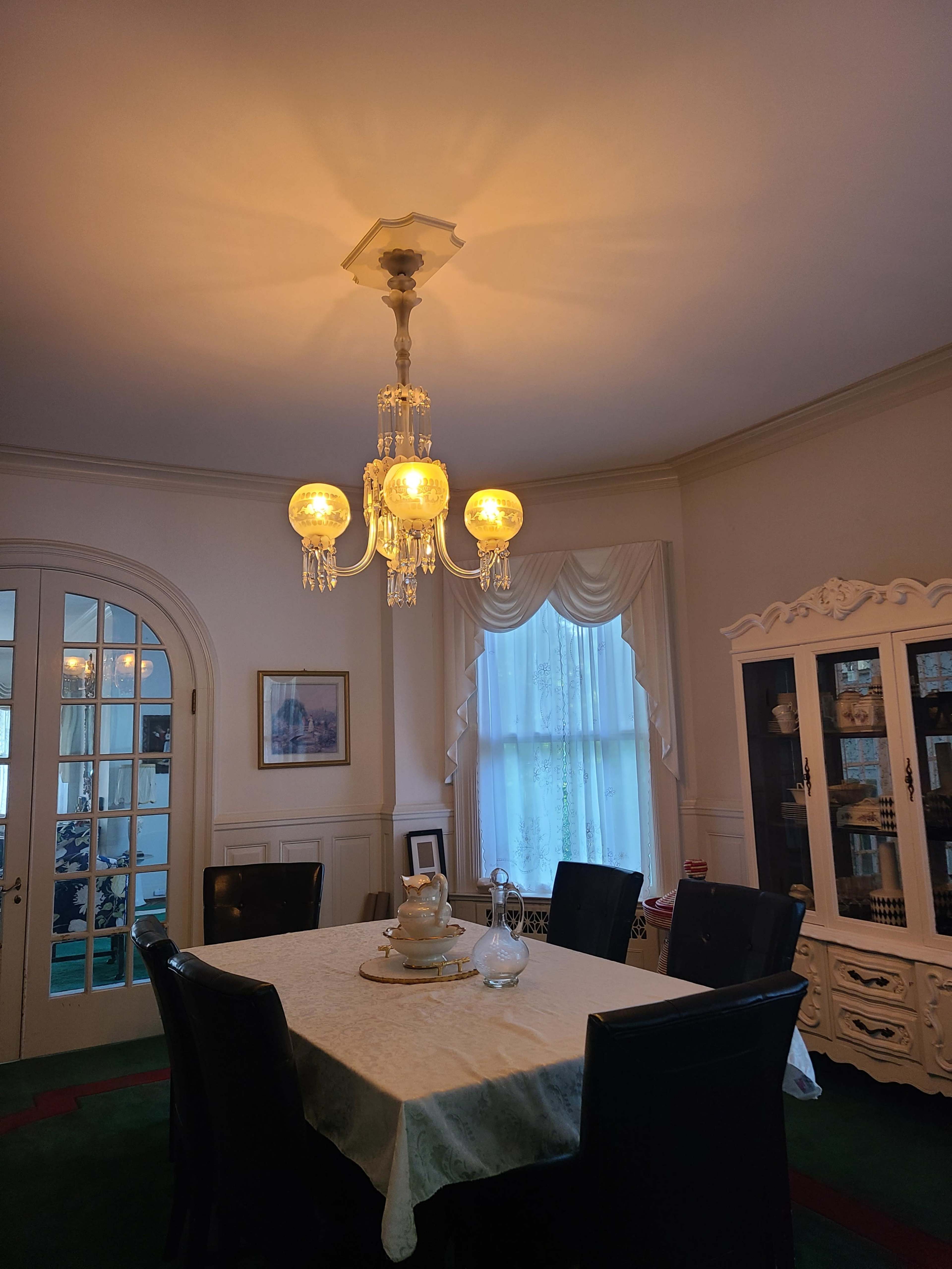 A dining room features a chandelier above a table set with a teapot and a glass pitcher, surrounded by dark chairs and illuminated by soft light.