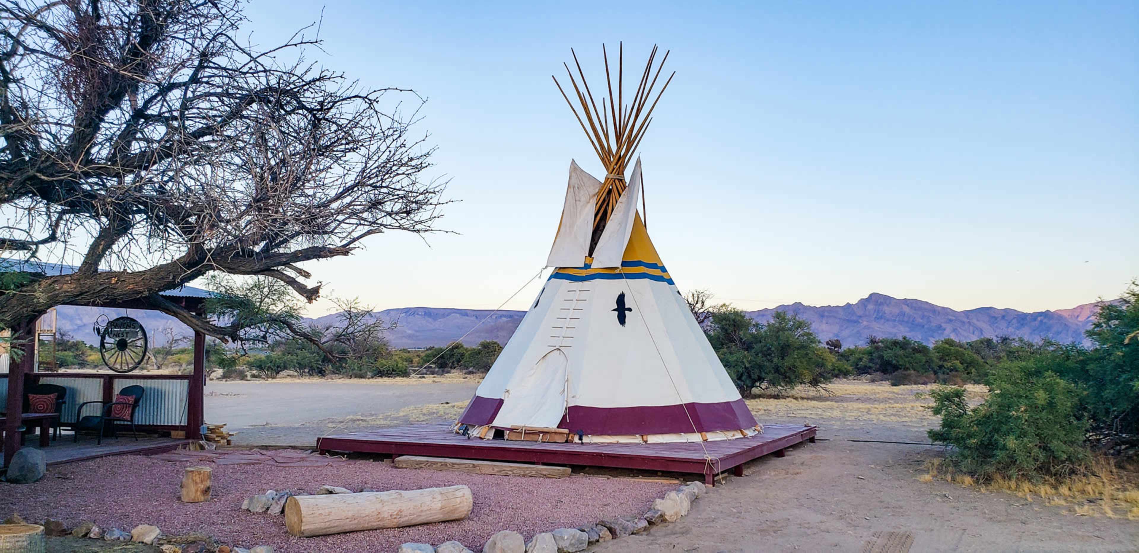 A teepee stands on a wooden platform surrounded by shrubs and distant mountains under a clear sky.