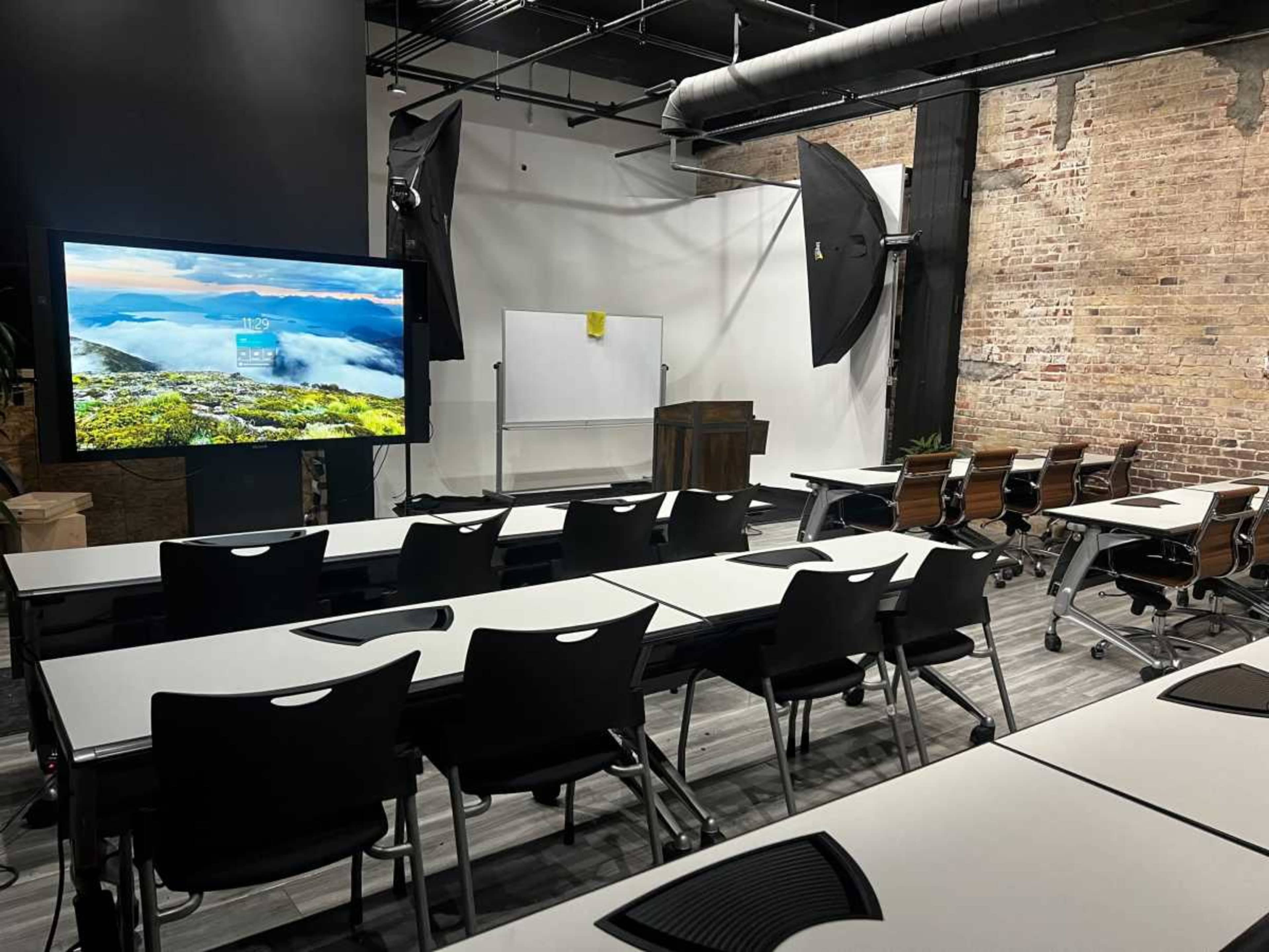 A classroom setup features rows of tables and chairs facing a large screen, with a whiteboard and a wooden podium at the front.