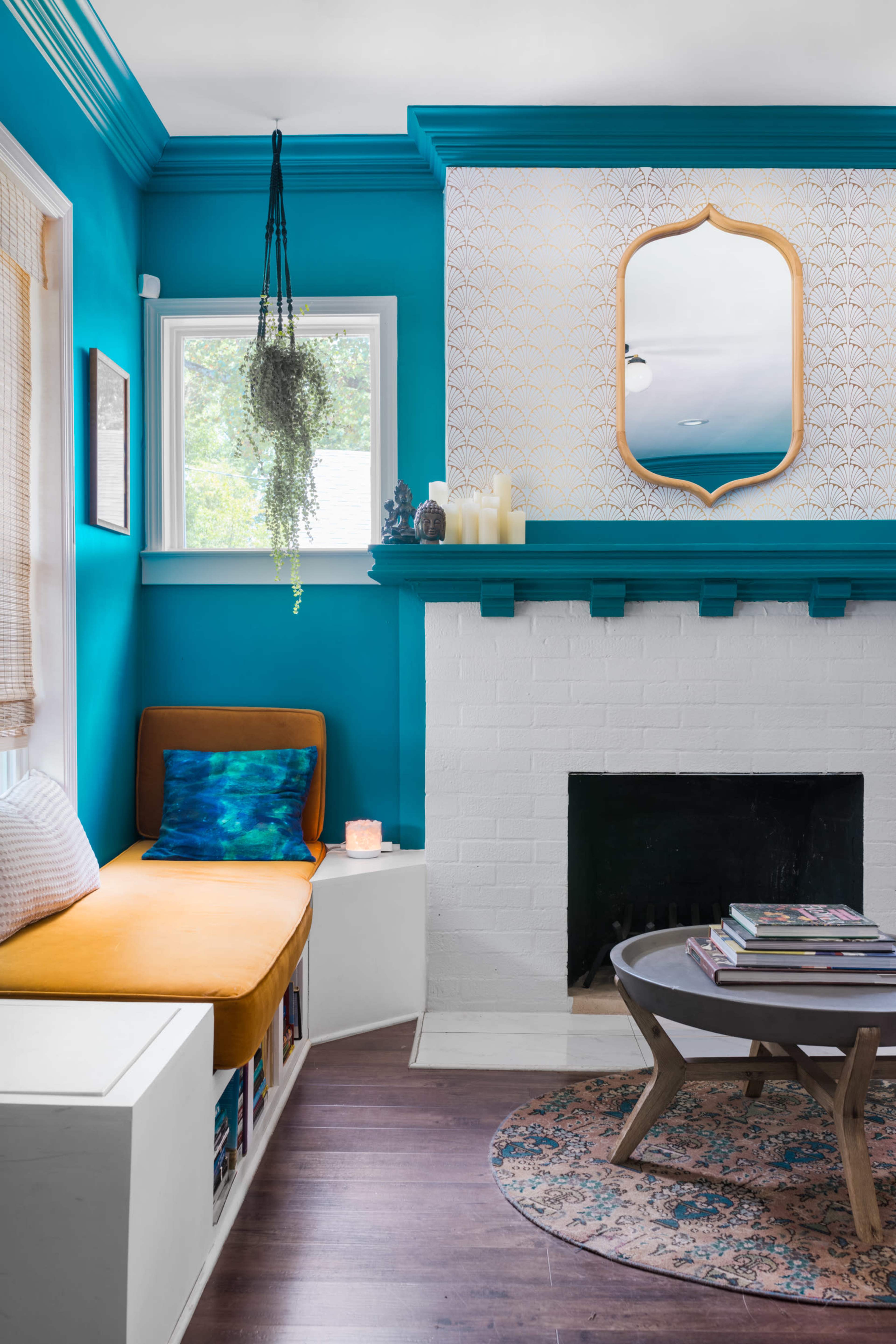 The image shows a bright room with turquoise walls, featuring a cozy seating area with an orange cushion, a white brick fireplace, and a round coffee table stacked with books.