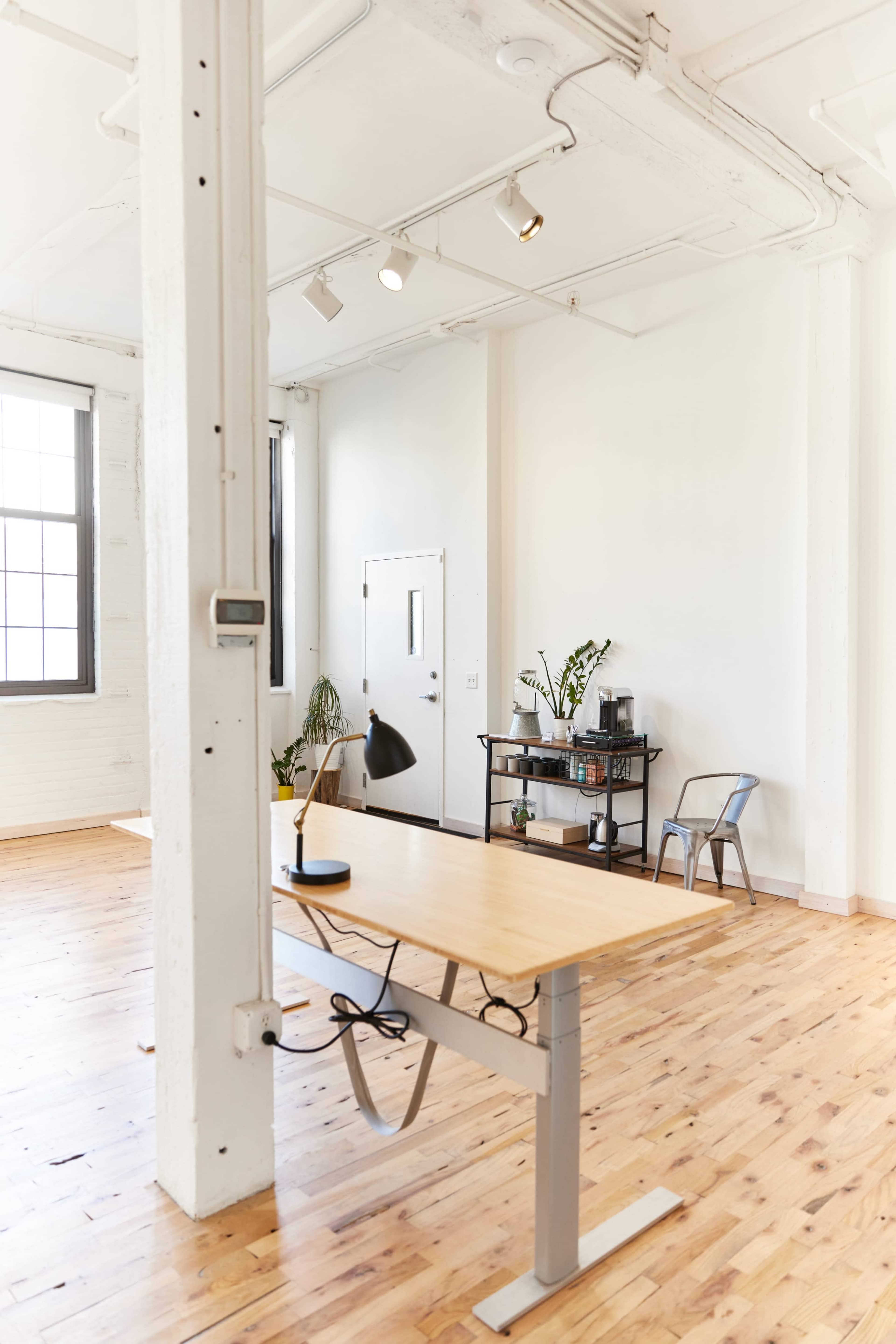 A minimalist office space with a wooden desk, a chair, and a plant against white walls and large windows.