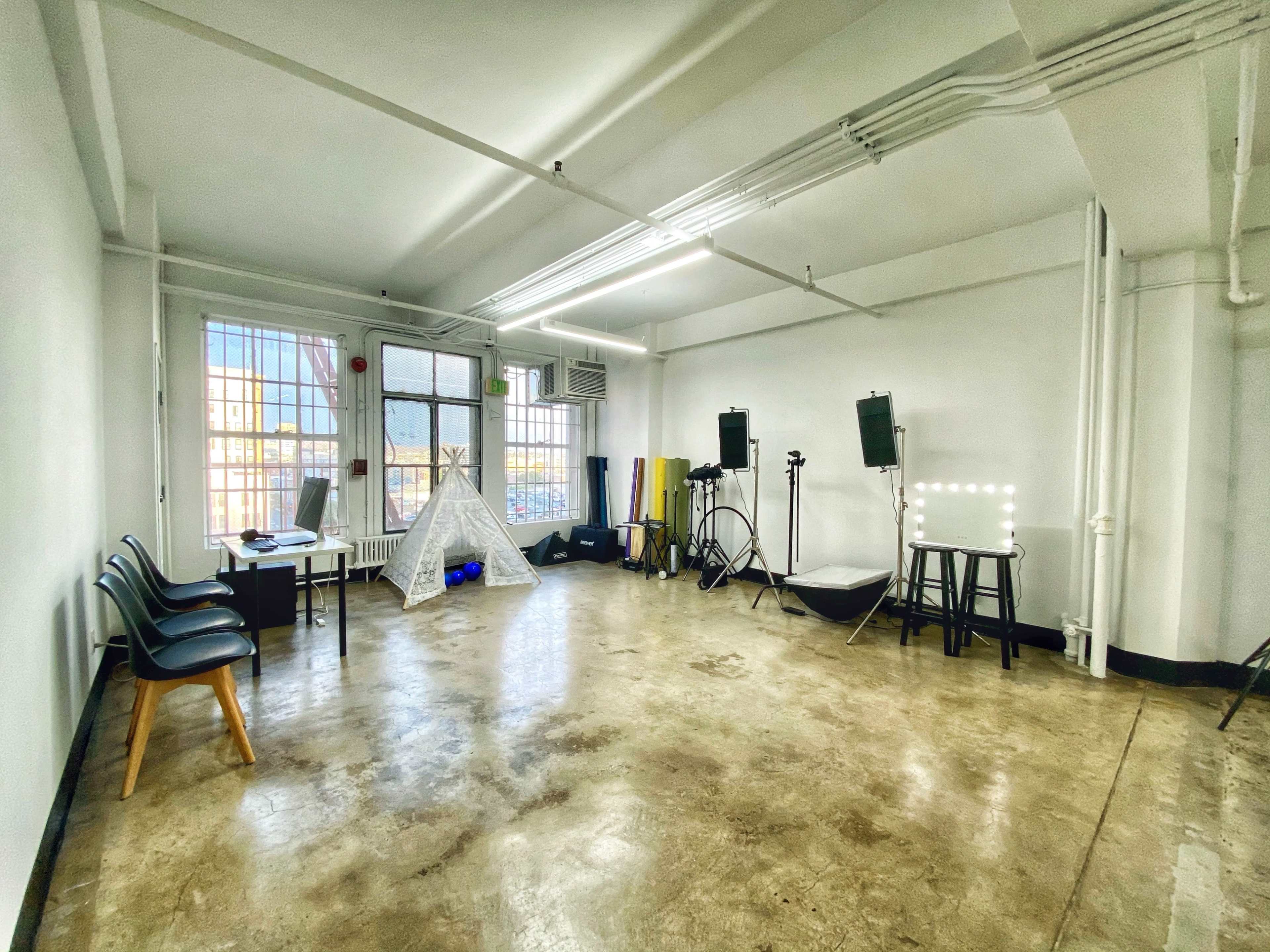 A spacious, empty studio with large windows, a desk with a laptop, and several lighting equipment setups along the walls.